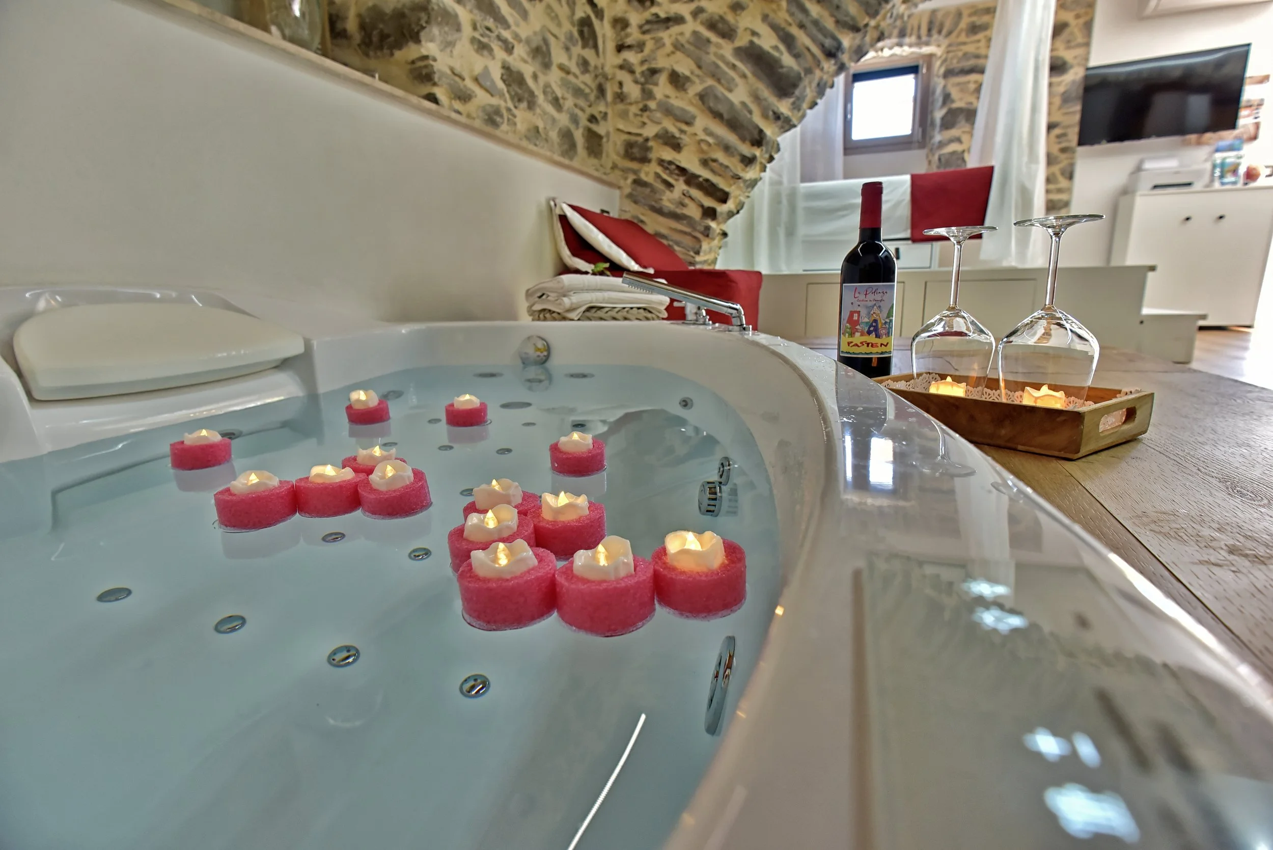 Vasca idromassaggio con candele galleggianti rosa e bianche, bottiglia di vino rosso, due bicchieri di vino vuoti, in una stanza con pareti in pietra e un divano rosso sullo sfondo