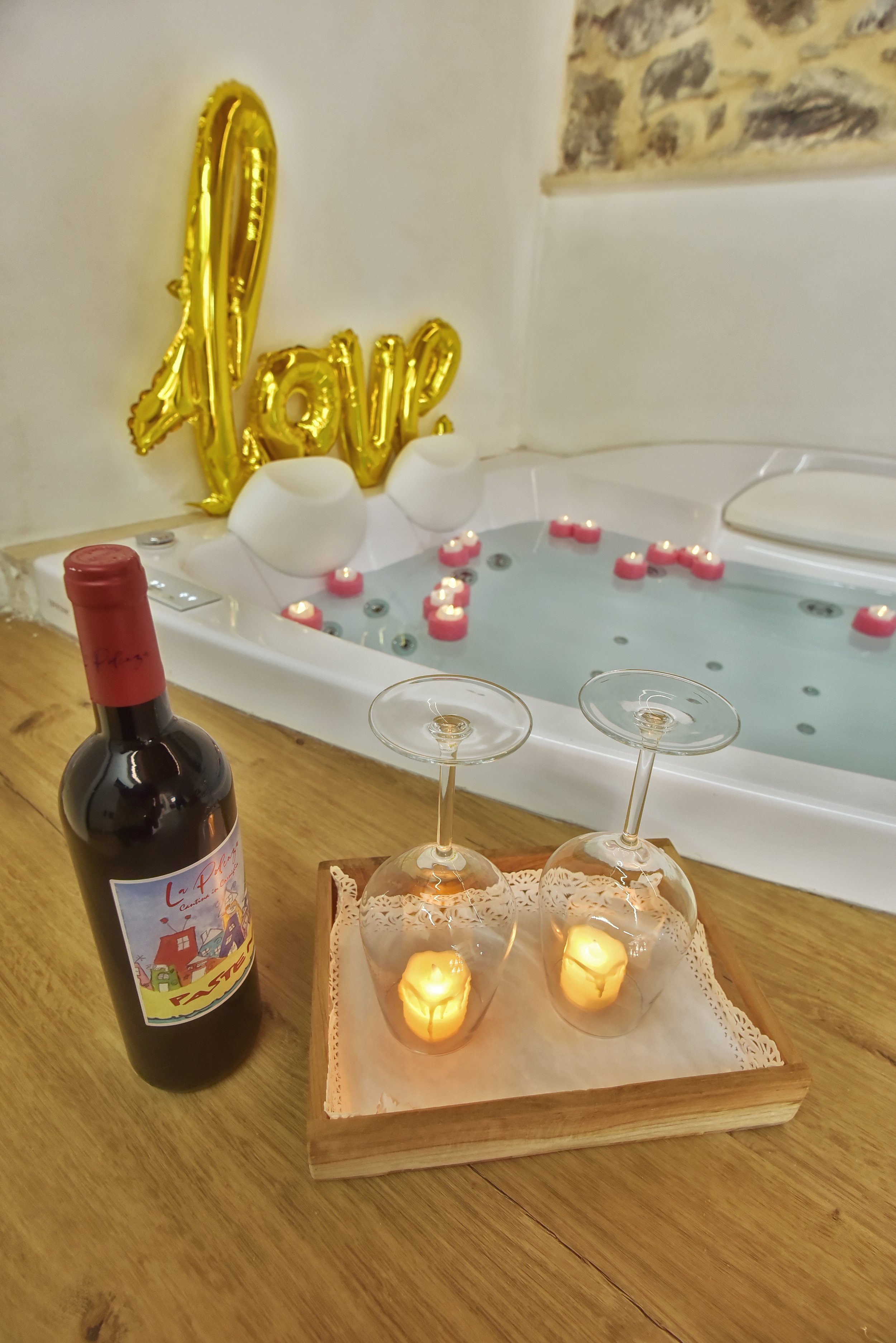 Decorazioni per una festa con un grande palloncino dorato che dice 'Love', candele accese, bicchieri di vino, una bottiglia di vino, una jacuzzi con candele galleggianti e petali di rosa, pareti di pietra e bianca.