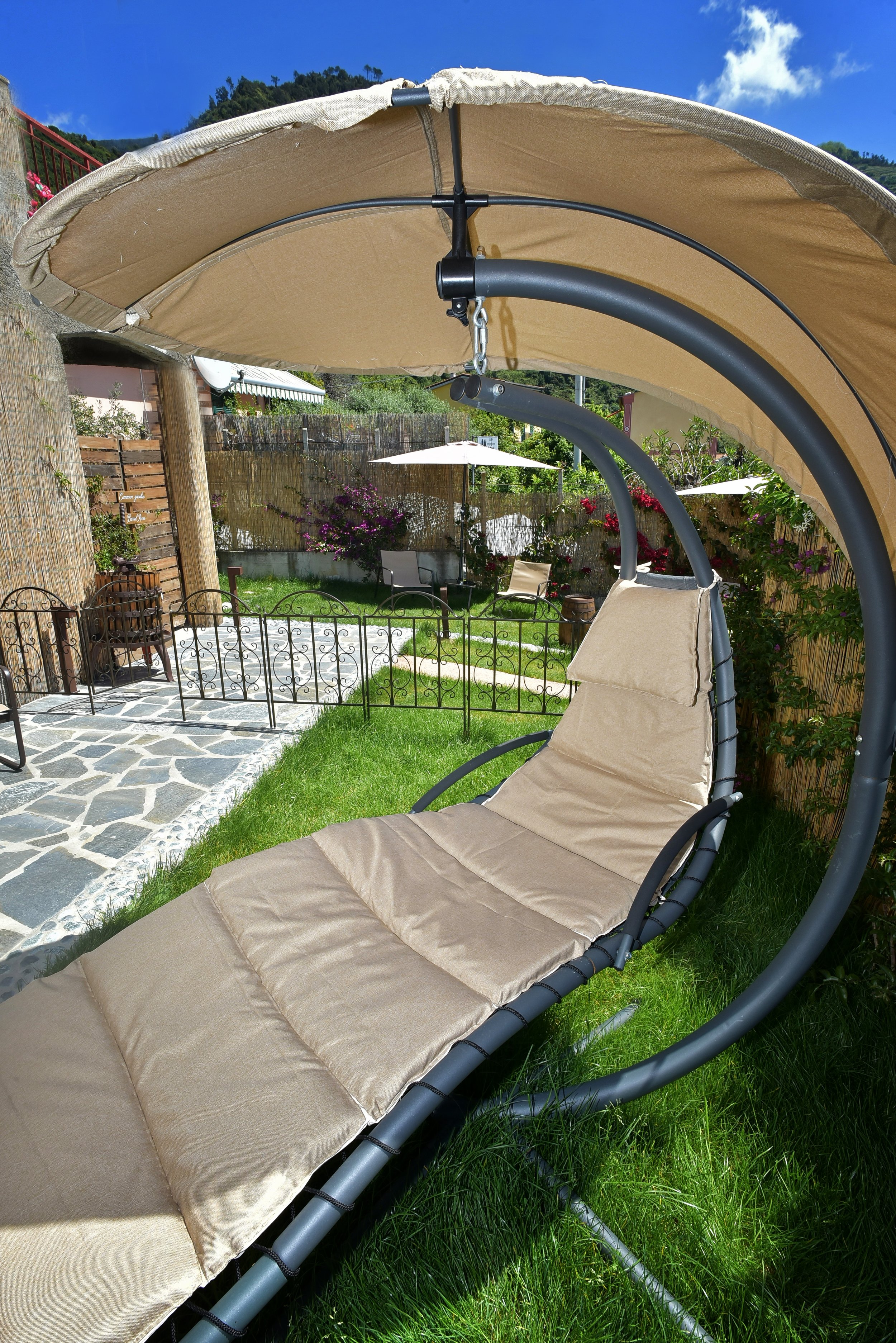 Sdraio con tettuccio beige in un giardino soleggiato, con piante, fiori e ombrelloni.