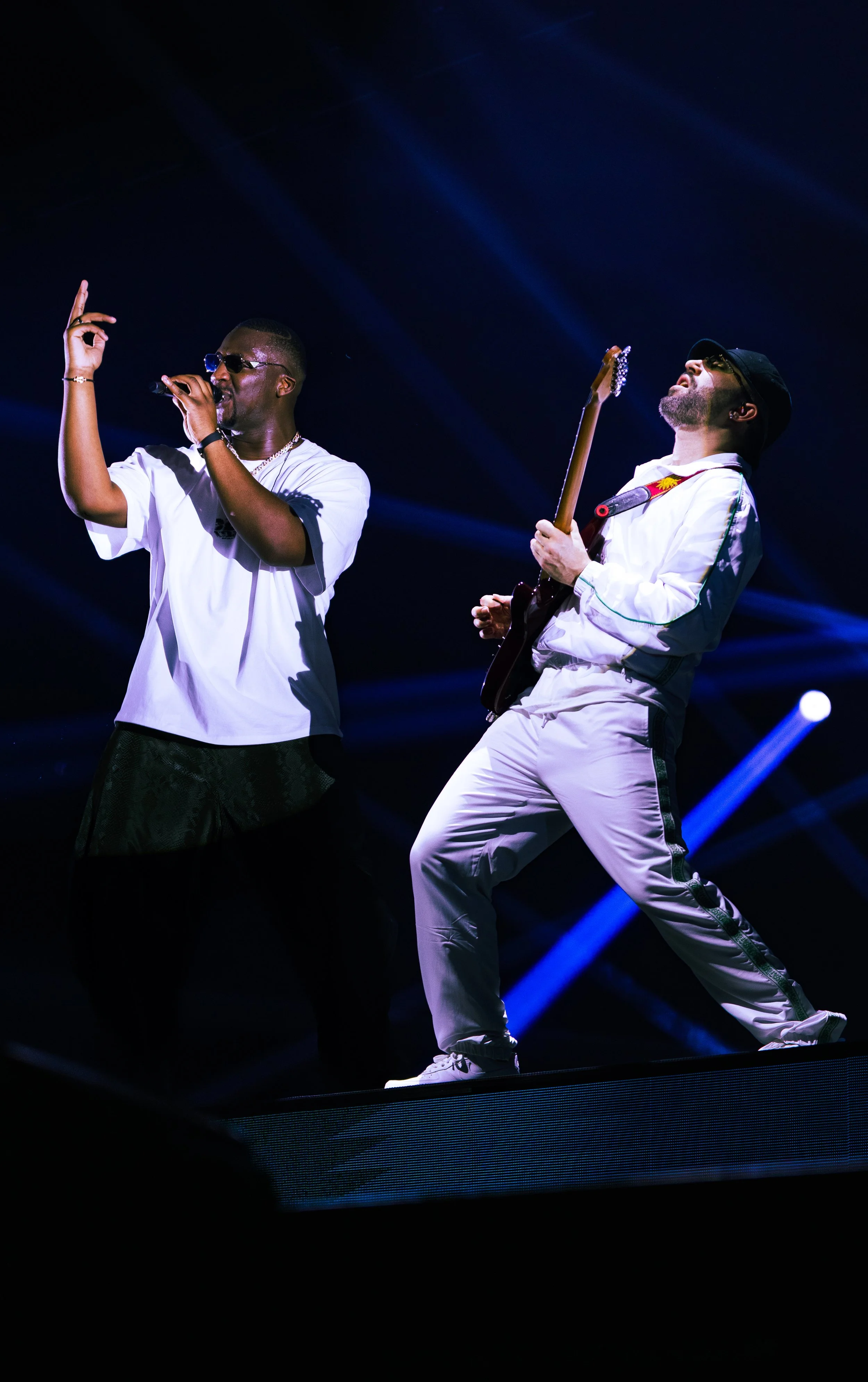 Deux musiciens en performance sur scène, un chanteur avec un micro et un guitariste portant des lunettes de soleil, vêtus de blanc, sous un éclairage bleu foncé.