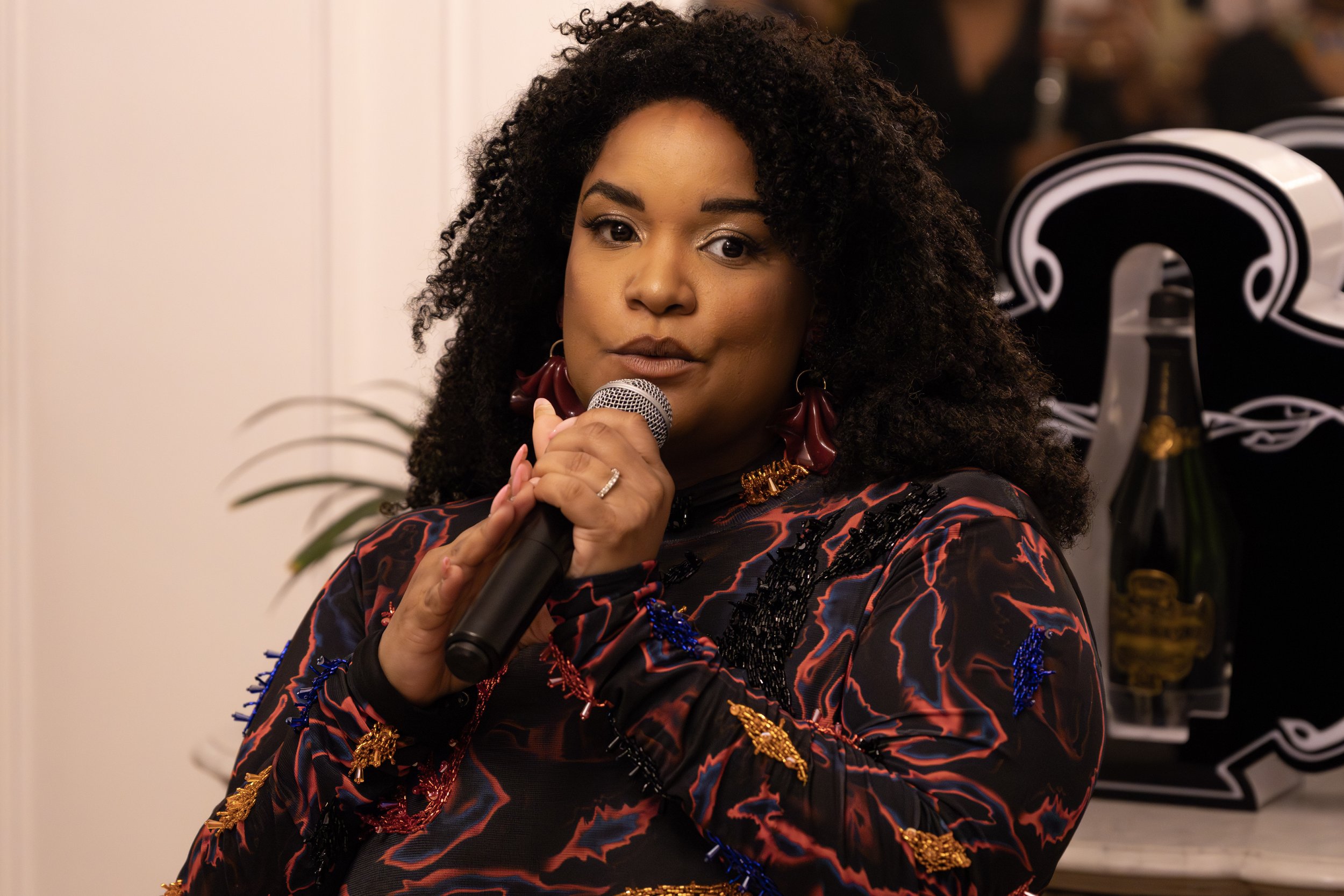 Une femme noire cheveux volumineux et bouclés tenant un microphone, portant une veste à motif rouge et noir, avec des boucles d'oreilles rouges, en train de parler ou de chanter.