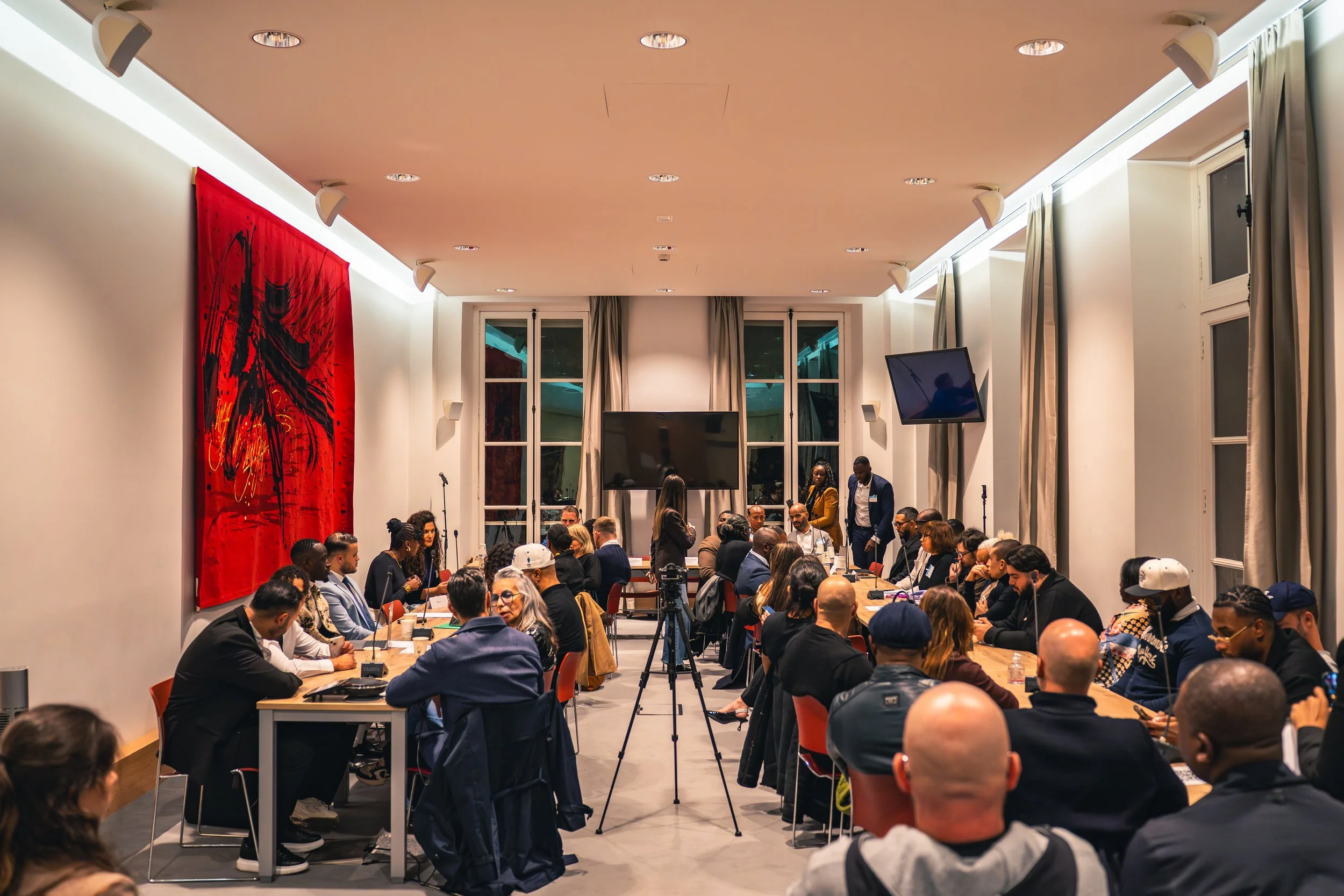 Une grande salle de réunion ou conférence remplie de personnes assises autour de longues tables, avec des écrans et des lampes suspendues au plafond. La pièce est lumineuse, avec des fenêtres en arrière-plan et des rideaux gris.
