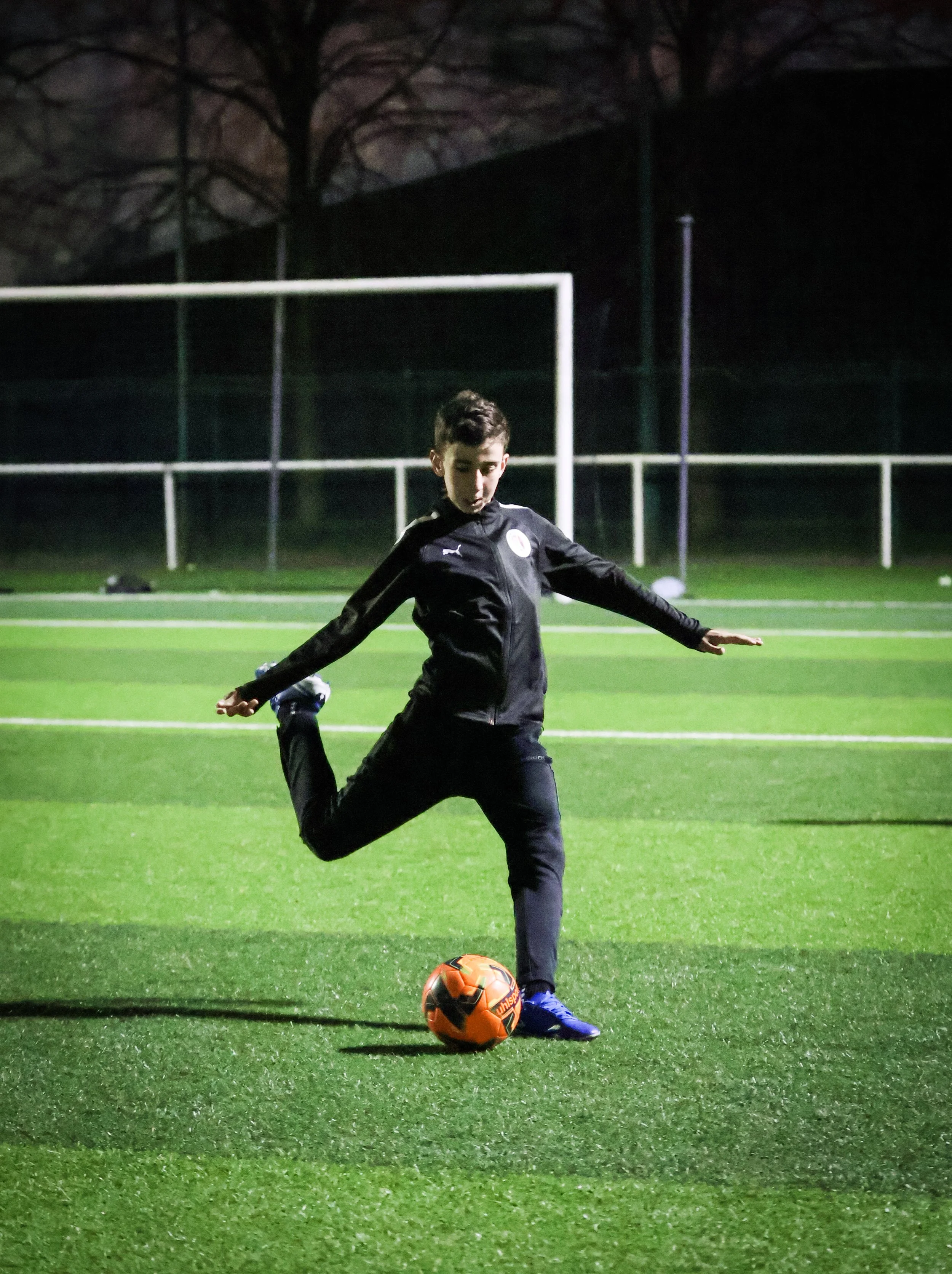 Un garçon en tenue de sport noire joue au football sur un terrain éclairé la nuit. Il s'apprête à frapper le ballon orange situé devant lui, avec un terrain de football en gazon synthétique vert et des buts en arrière-plan.