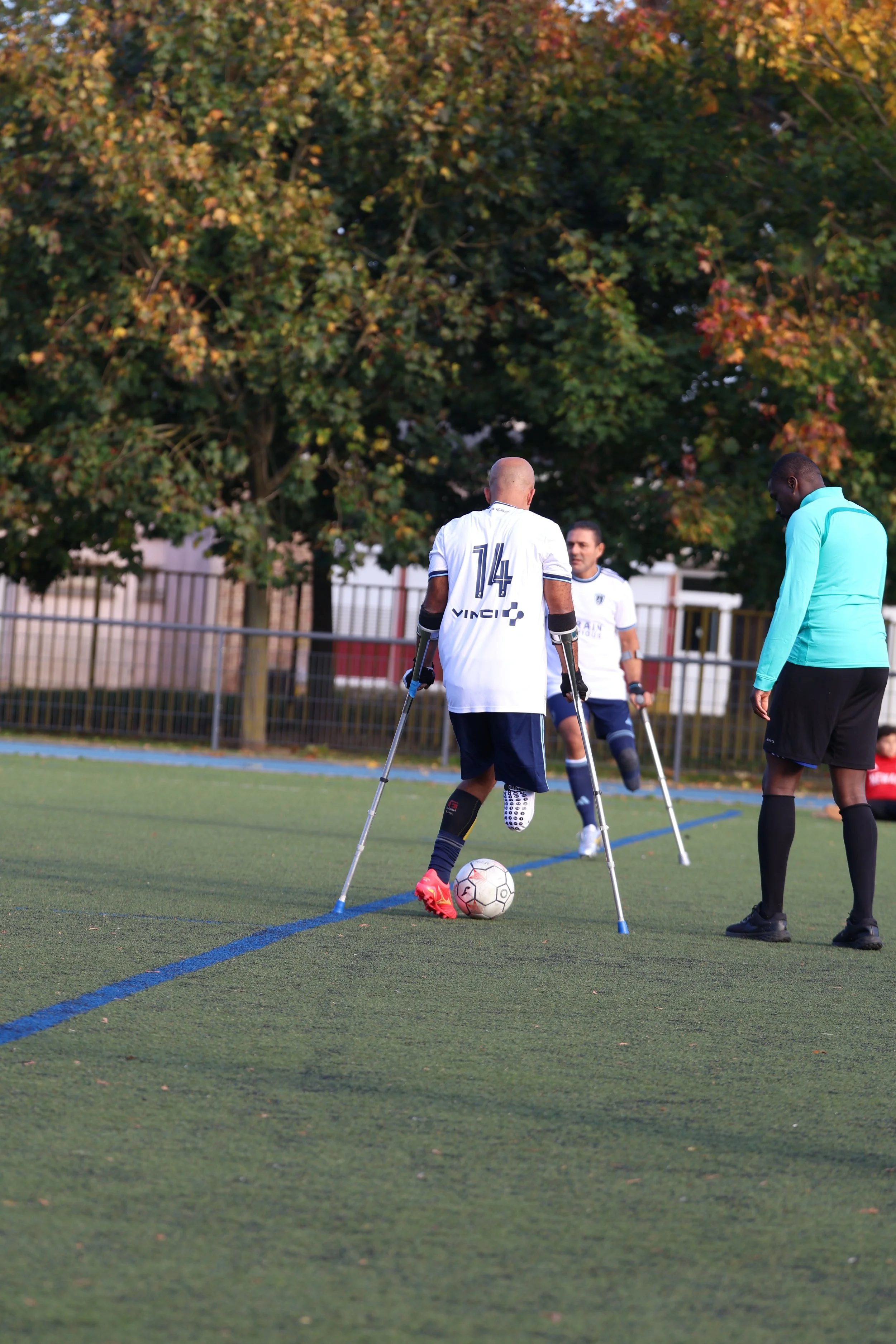 Trois hommes sur un terrain de sport, un d'entre eux avec des béquilles, portant un maillot blanc, entraînent la balle de football, une personne porte une veste bleue.