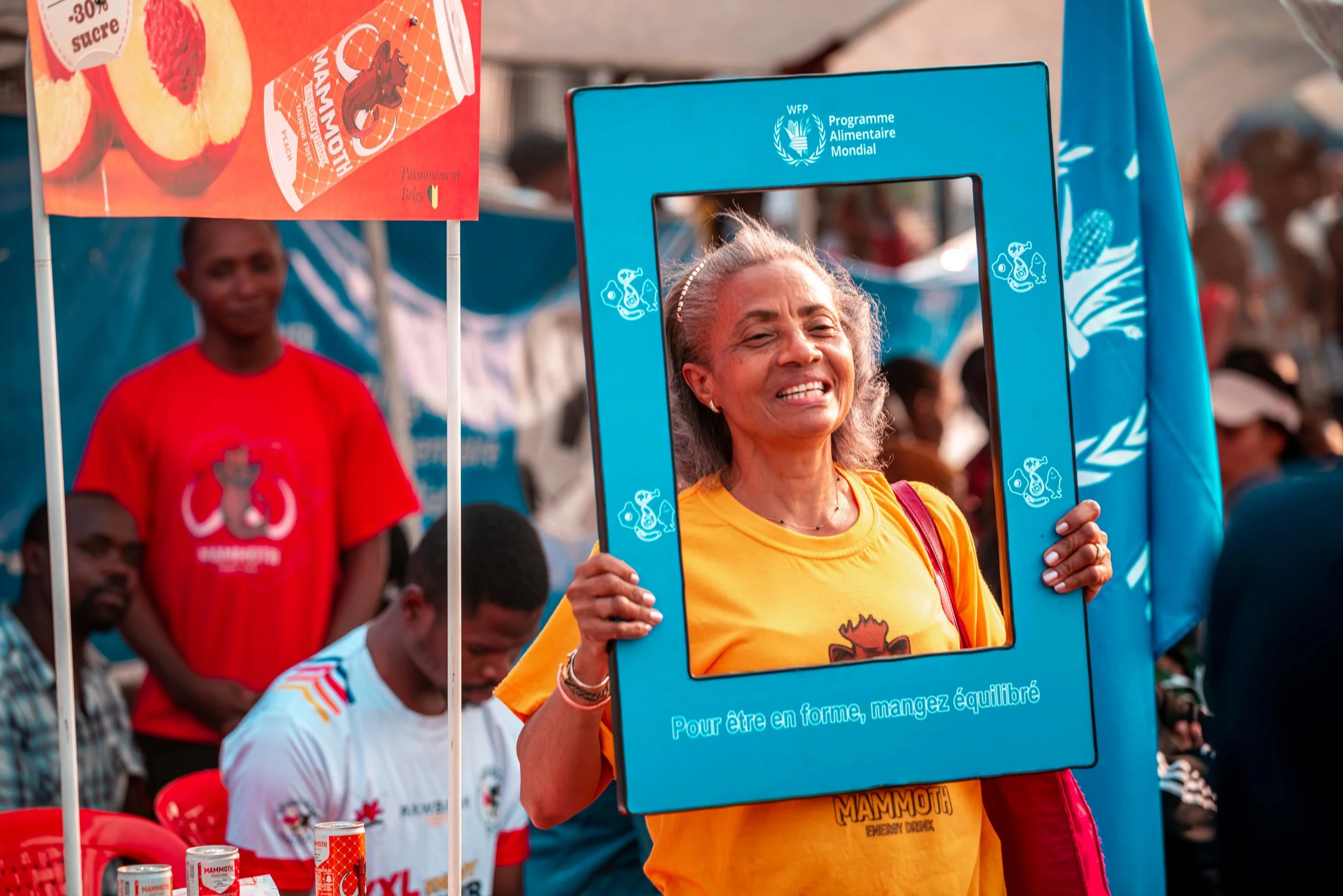 Une femme souriante tient un cadre bleu représentant une personne derrière une affiche promotionnelle lors d'un événement public. En arrière-plan, plusieurs personnes assistent à l'événement, et il y a des affiches et des produits Mammoth sur une table.