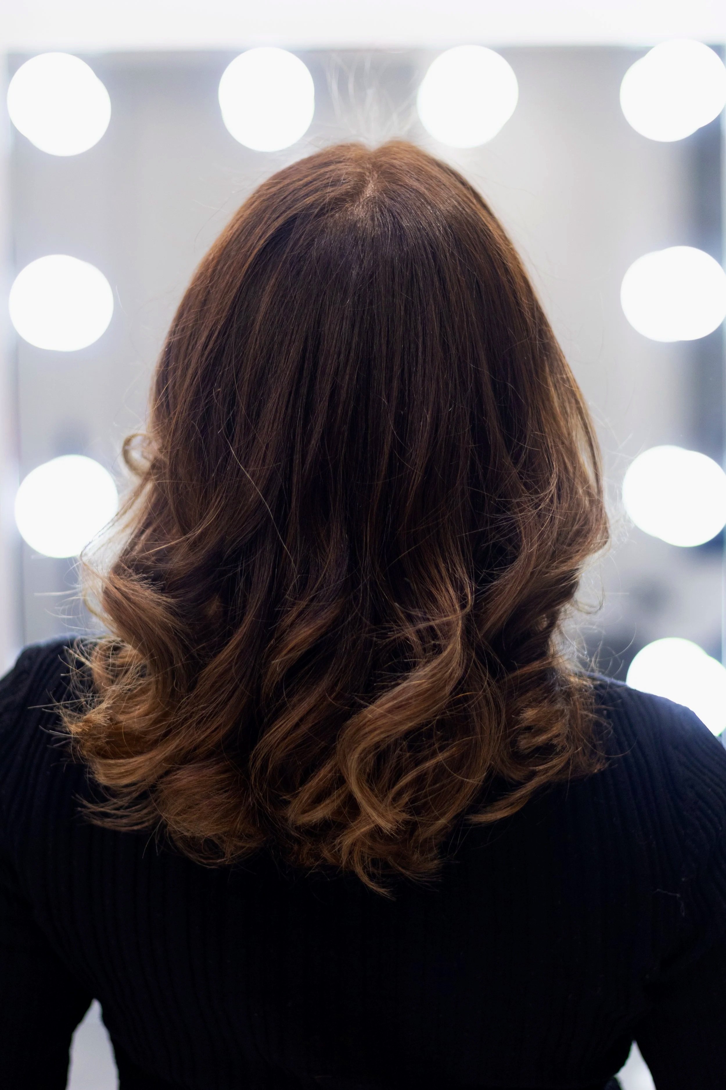 Une femme vue de dos avec de longs cheveux bouclés bruns portant un pull noir, devant un miroir éclairé par des ampoules.