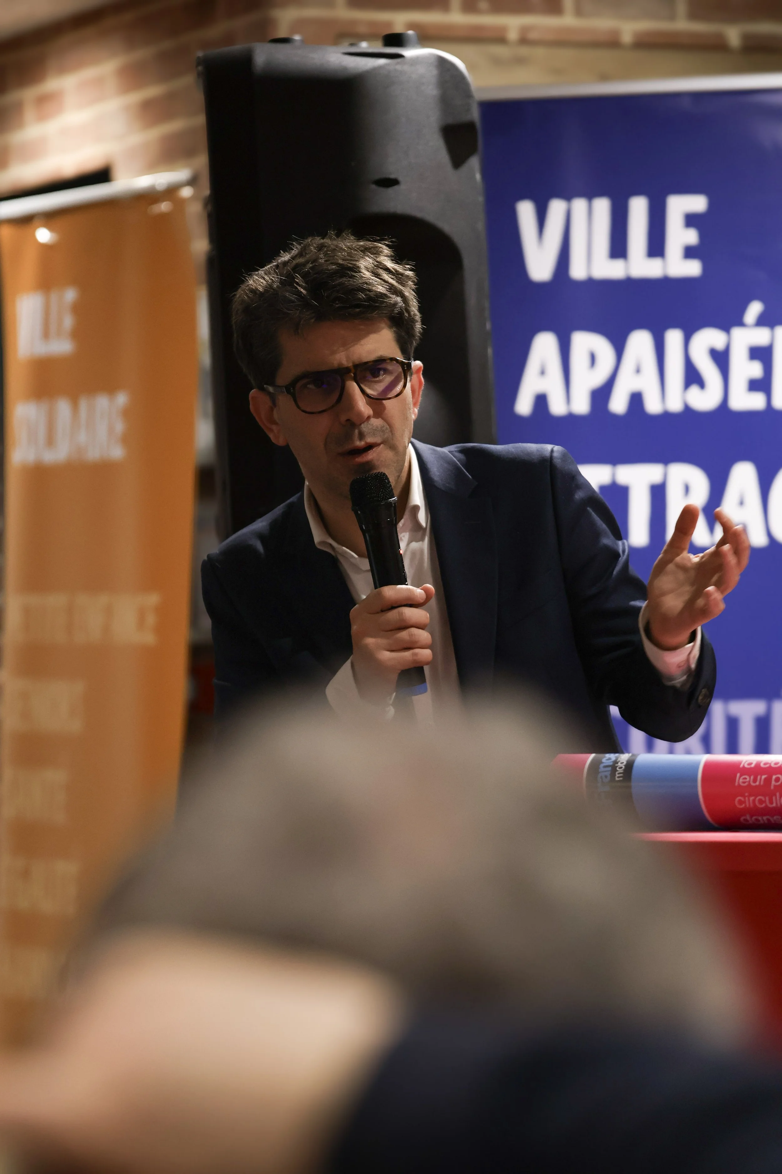Un homme en costume parle en tenant un microphone lors d'une présentation. En arrière-plan, un panneau bleu affiche le texte "VILLE" et d'autres phrases. À gauche, une bannière orange est partiellement visible.