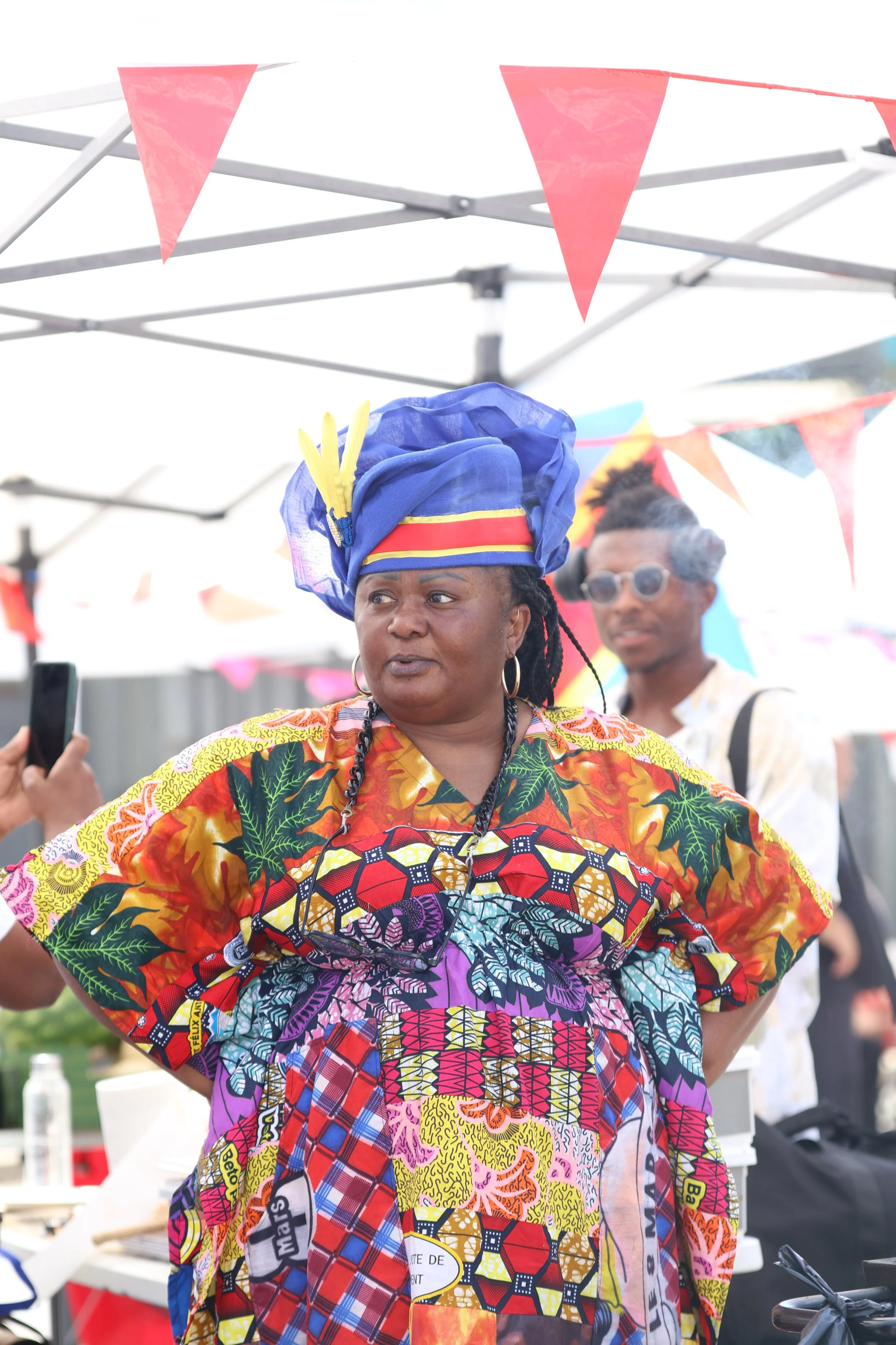 Une femme portant une robe africaine colorée et un chapeau bleu avec des décorations jaunes, se tenant devant un stand lors d'un événement en plein air, avec d'autres personnes et des décorations en fond.