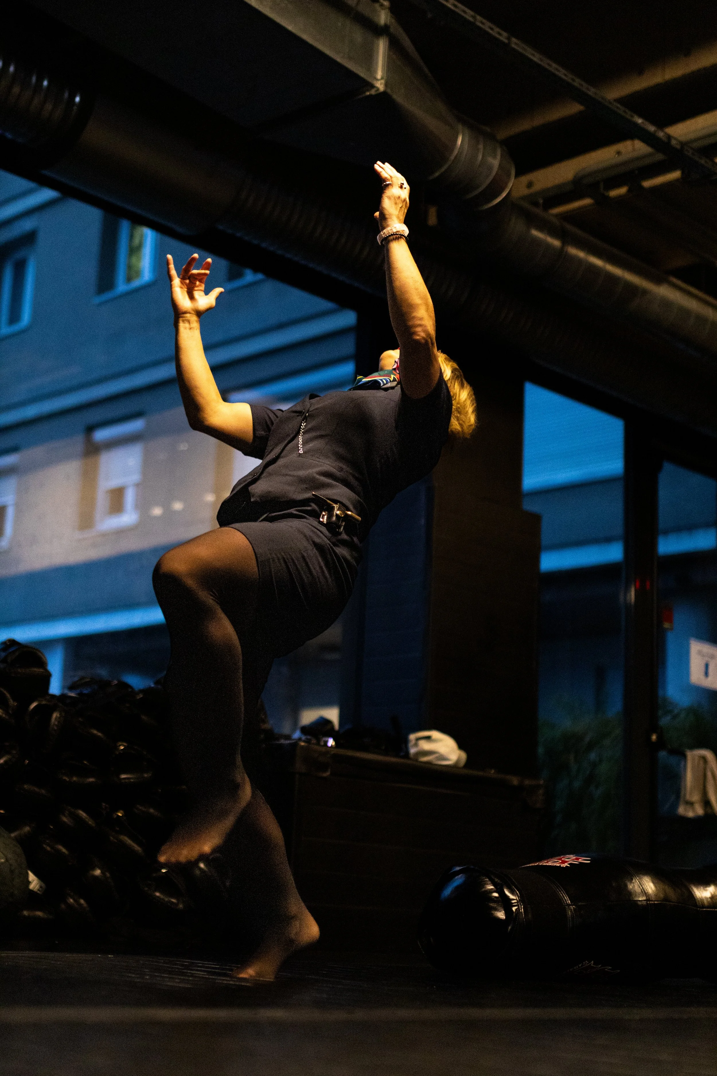 Une femme effectuant une posture de yoga ou de méditation en intérieur, avec des sacs de sport et un mur en arrière-plan.