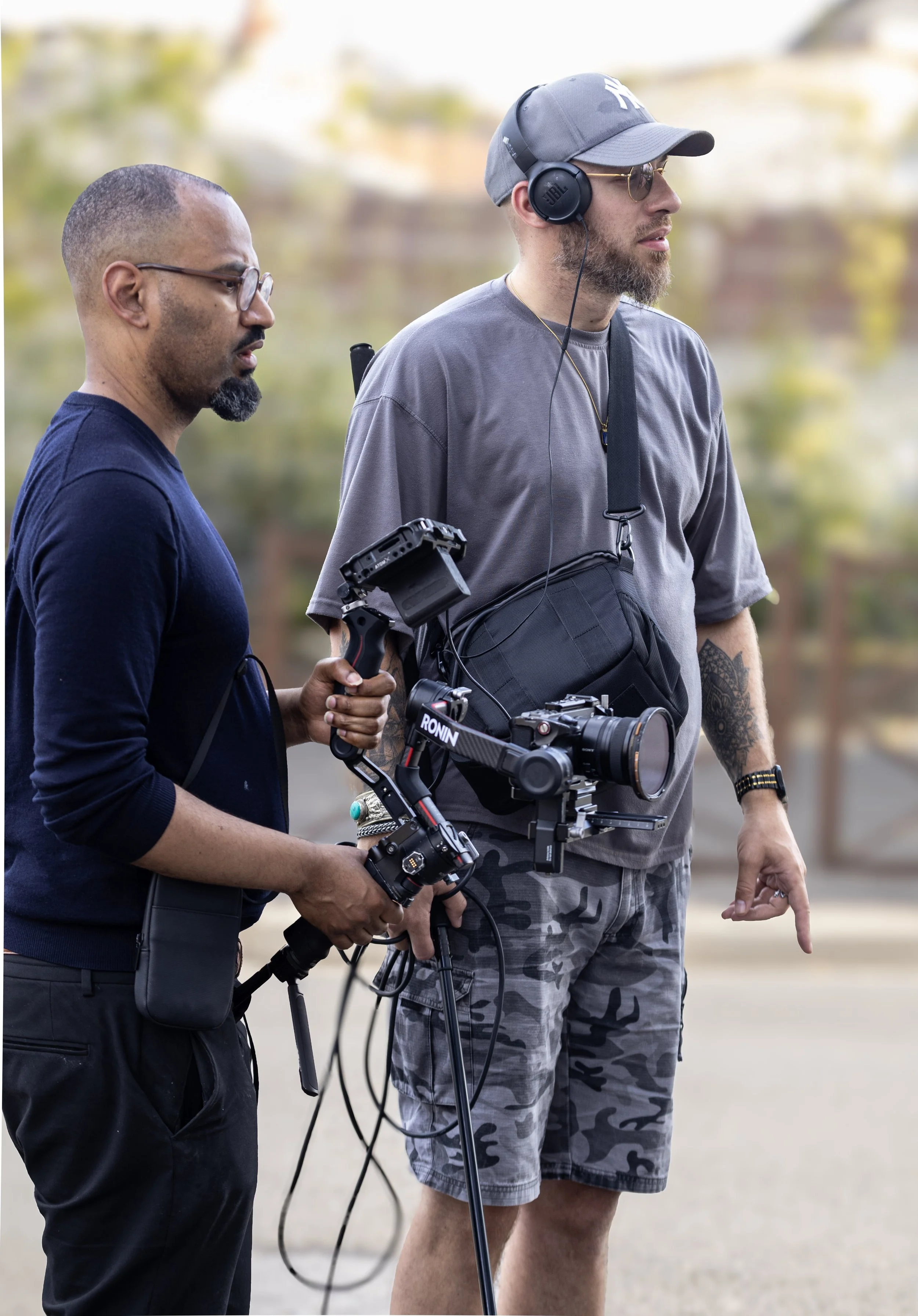 Deux hommes munis d'équipements de tournage vidéo discutent à l'extérieur, l'un tient une caméra stabilisée et l'autre porte un casque audio et une caméra, avec un fond flou de nature.