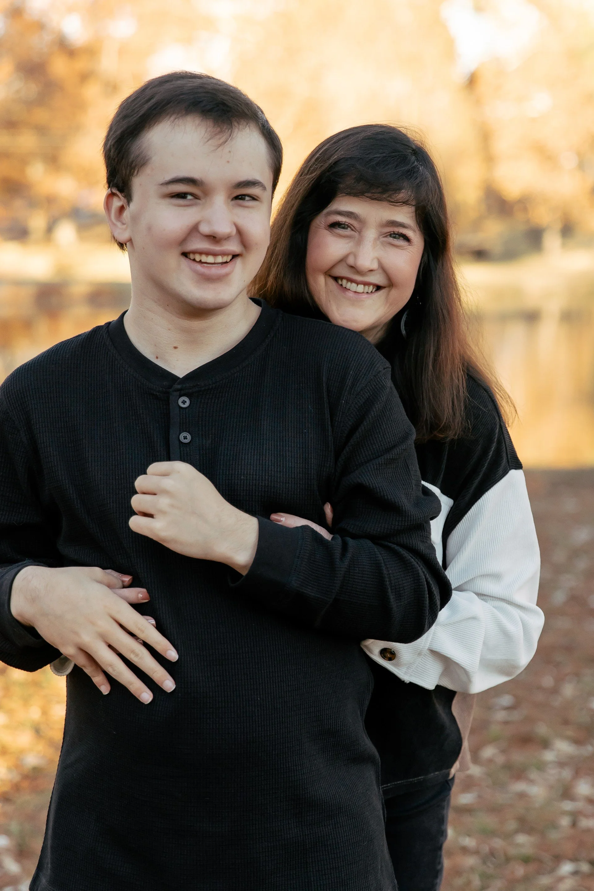 A young man and a woman are smiling and standing close together outdoors with autumn foliage in the background.