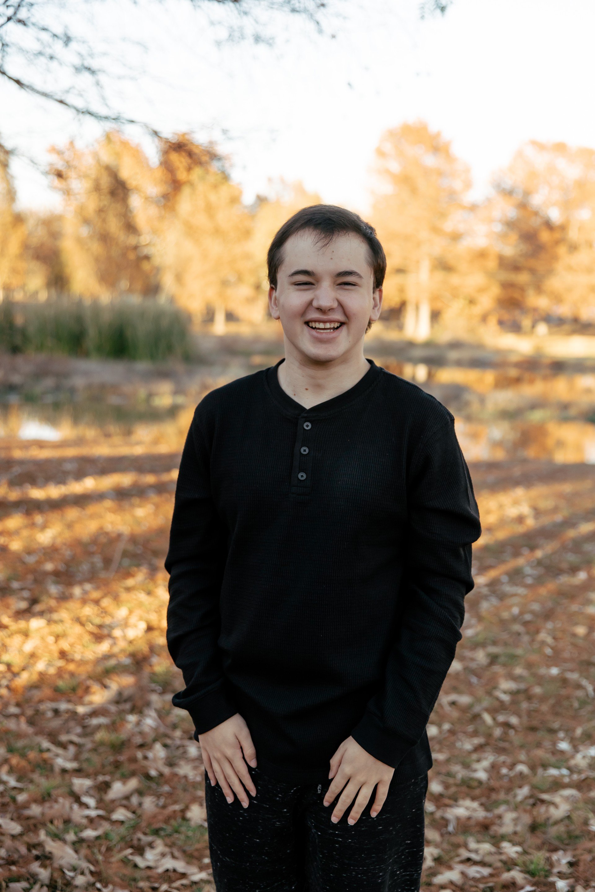 A young person with short dark hair smiling outdoors during autumn, wearing a black long-sleeve top and patterned pants, with trees and fallen leaves in the background.