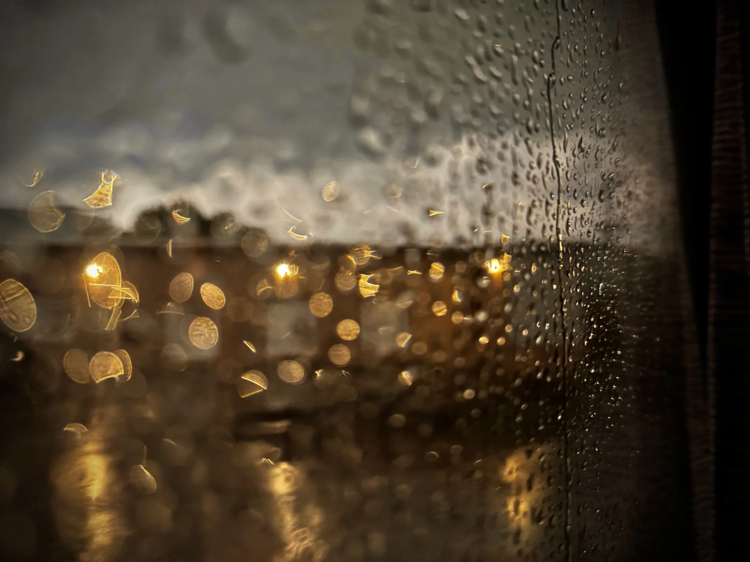 Rain droplets on a glass surface with blurry streetlights and cloudy sky in the background at dusk.