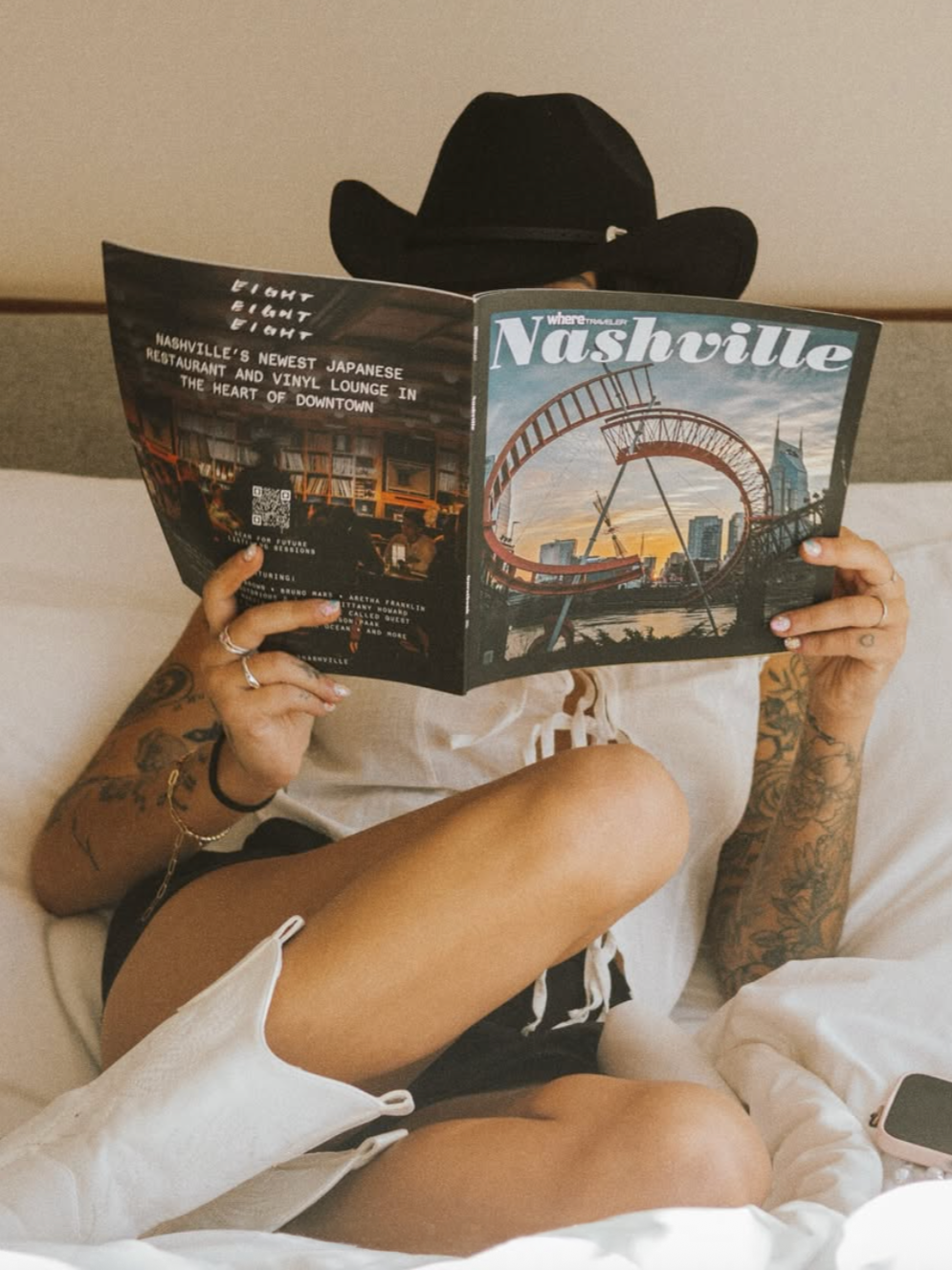 Person wearing a black hat, white shirt, and black shorts, with visible tattoos, sitting on a bed, reading a Nashville travel brochure.
