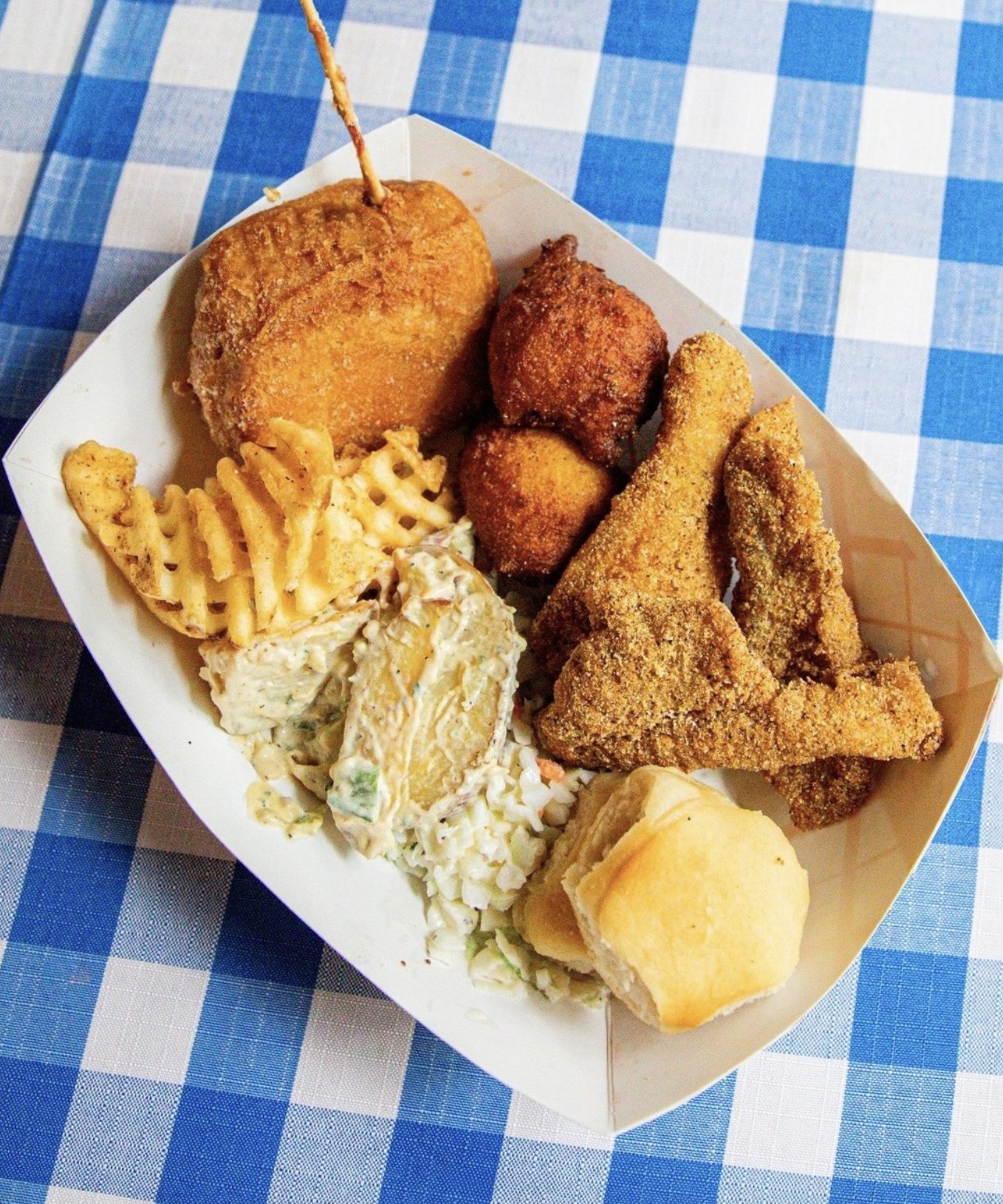 A paper tray of fried chicken, macaroni and cheese, coleslaw, potato salad, and roll on a blue checkered tablecloth.