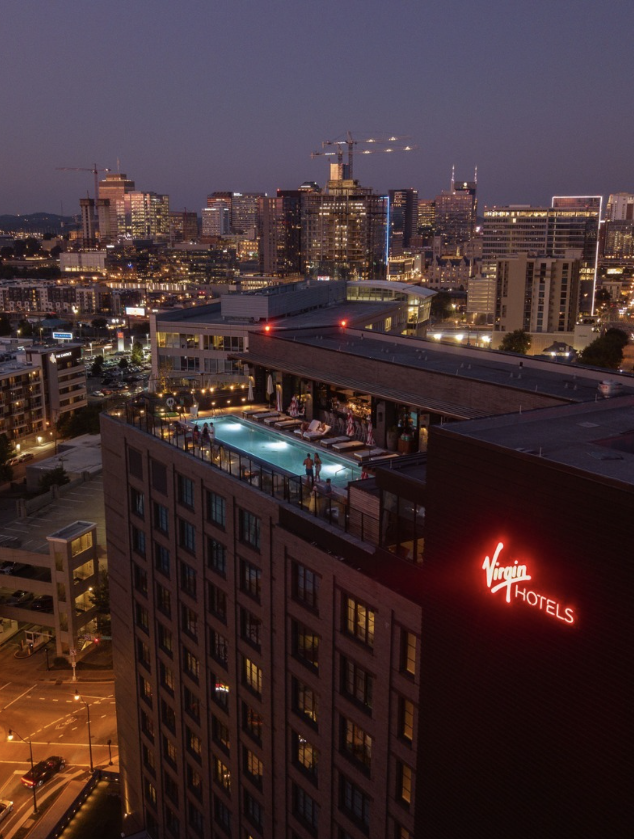 Nighttime cityscape view from an apartment building with a rooftop pool and a neon 'Virgin Hotels' sign on the building in the foreground. The skyline is illuminated with numerous lit windows and cranes.