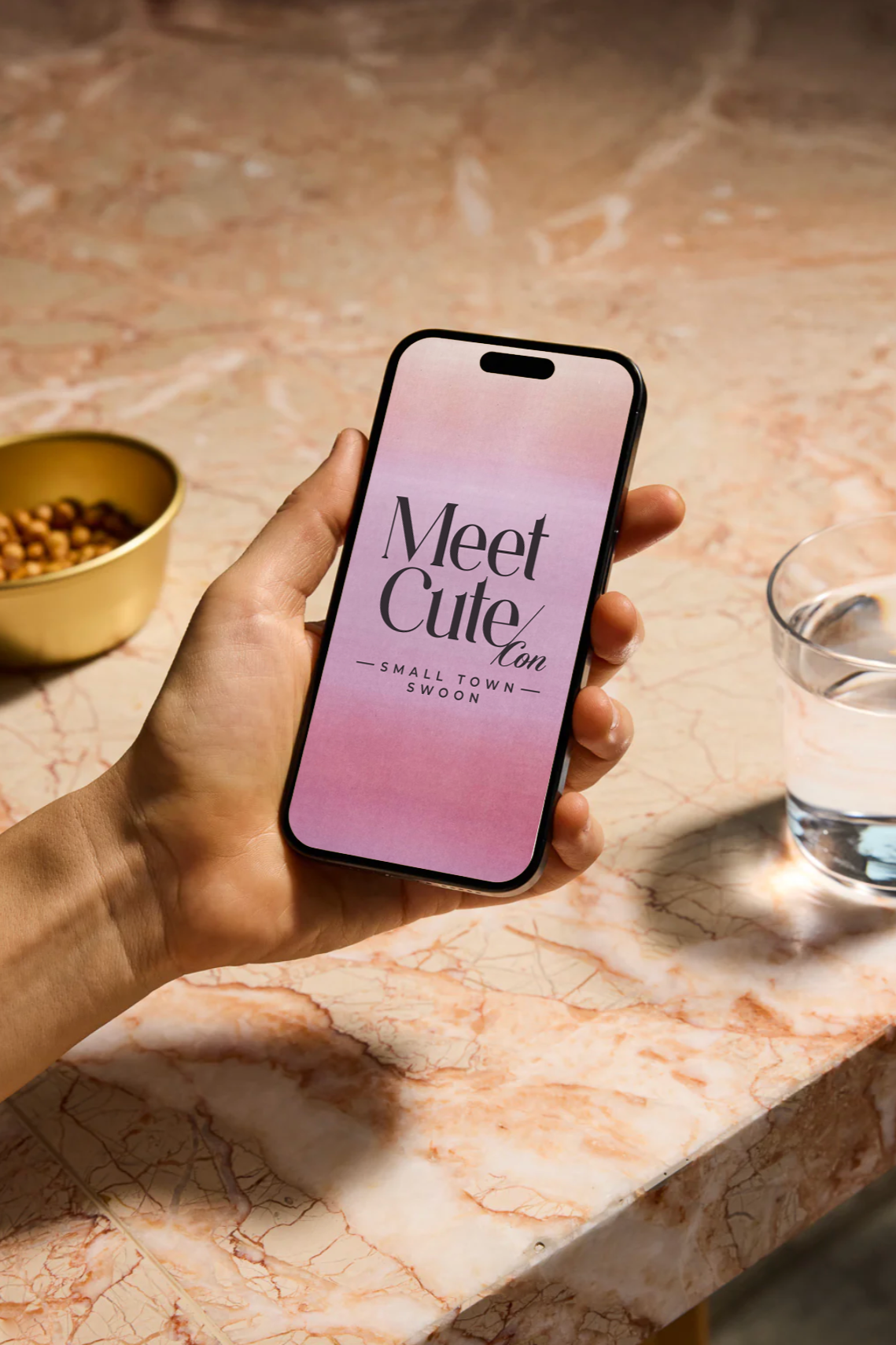 A person holding a smartphone displaying a dating app that says 'Meet Cute con Small Town Swoon'. The background features a marble countertop, a bowl of snacks on the left, and a glass of water on the right.