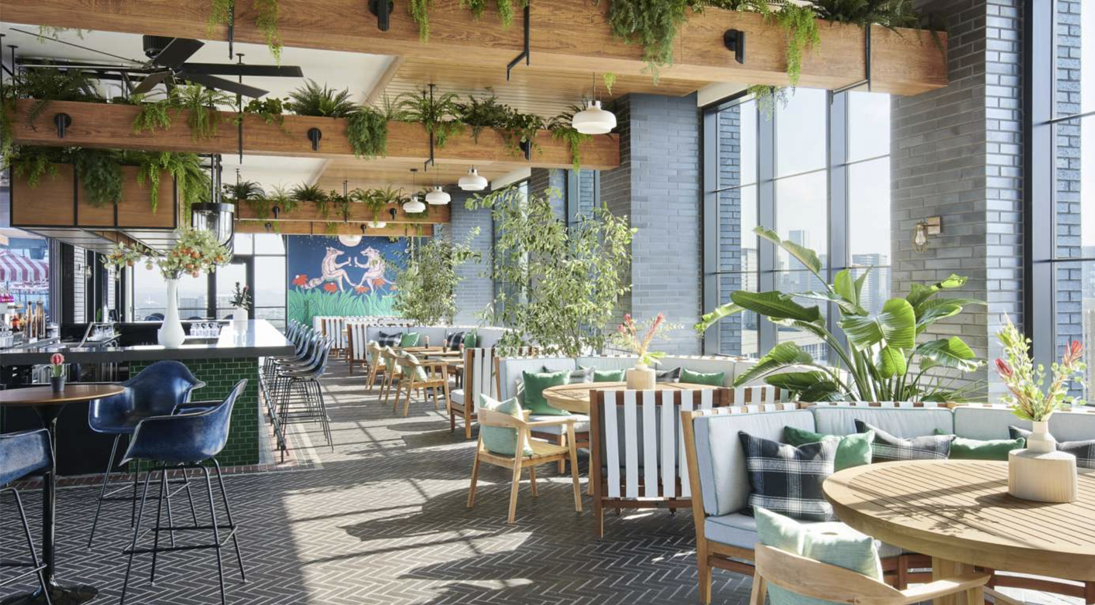 Bright indoor restaurant with large windows, green plants, wooden accents, and colorful artwork featuring two unicorns and mushrooms.