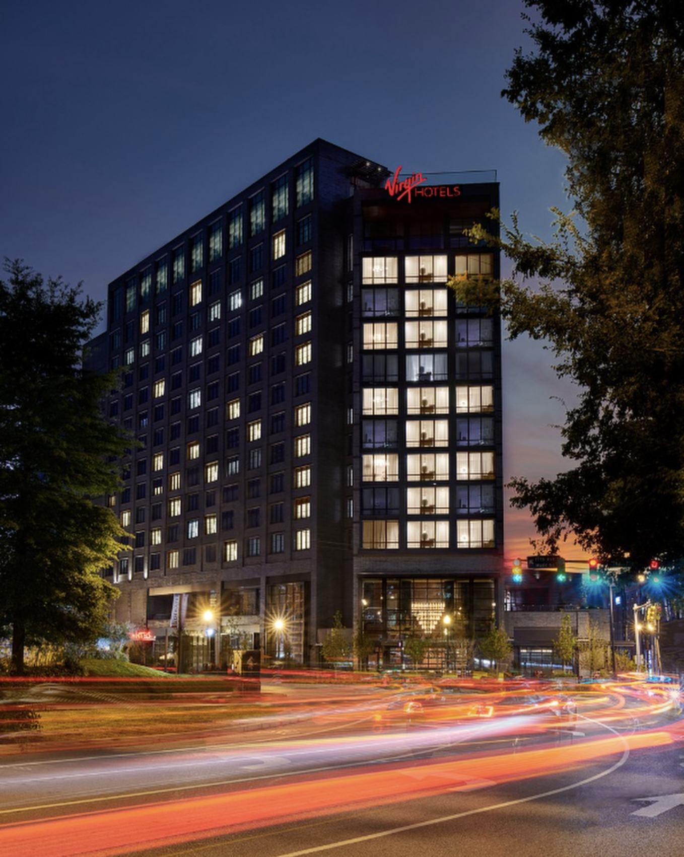 Nighttime photo of a tall hotel building with illuminated windows and the red Virginia Hotels sign on top. Light trails from moving vehicles are visible on the street in front of the building, with trees on either side.