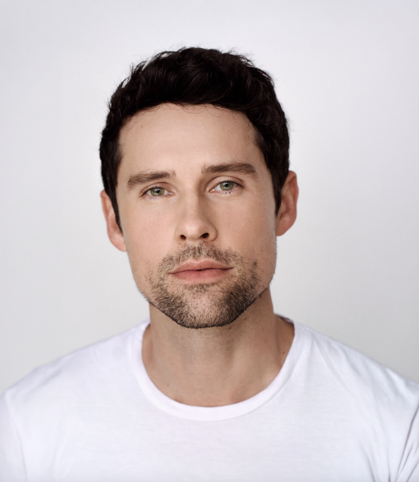 Close-up portrait of a young man with dark wavy hair, light skin, blue-green eyes, and a light beard, wearing a white t-shirt against a plain light background.