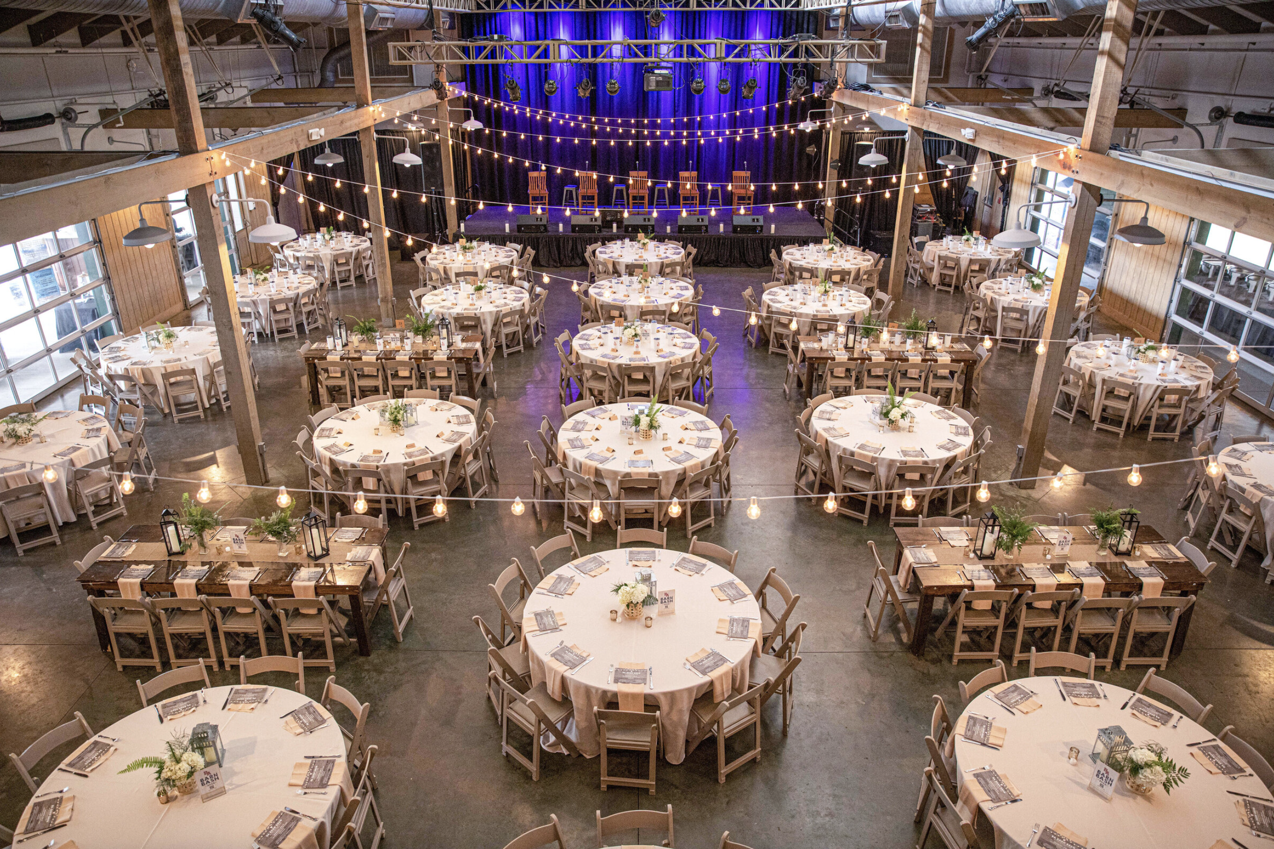 An indoor event space decorated with round tables covered in beige tablecloths, adorned with small flower arrangements, set for a celebration. Overhead, string lights hang across the space, and a stage with a blue curtain and five wooden chairs is at the far end. The room features large windows along one side, and the ceiling is supported by wooden beams.