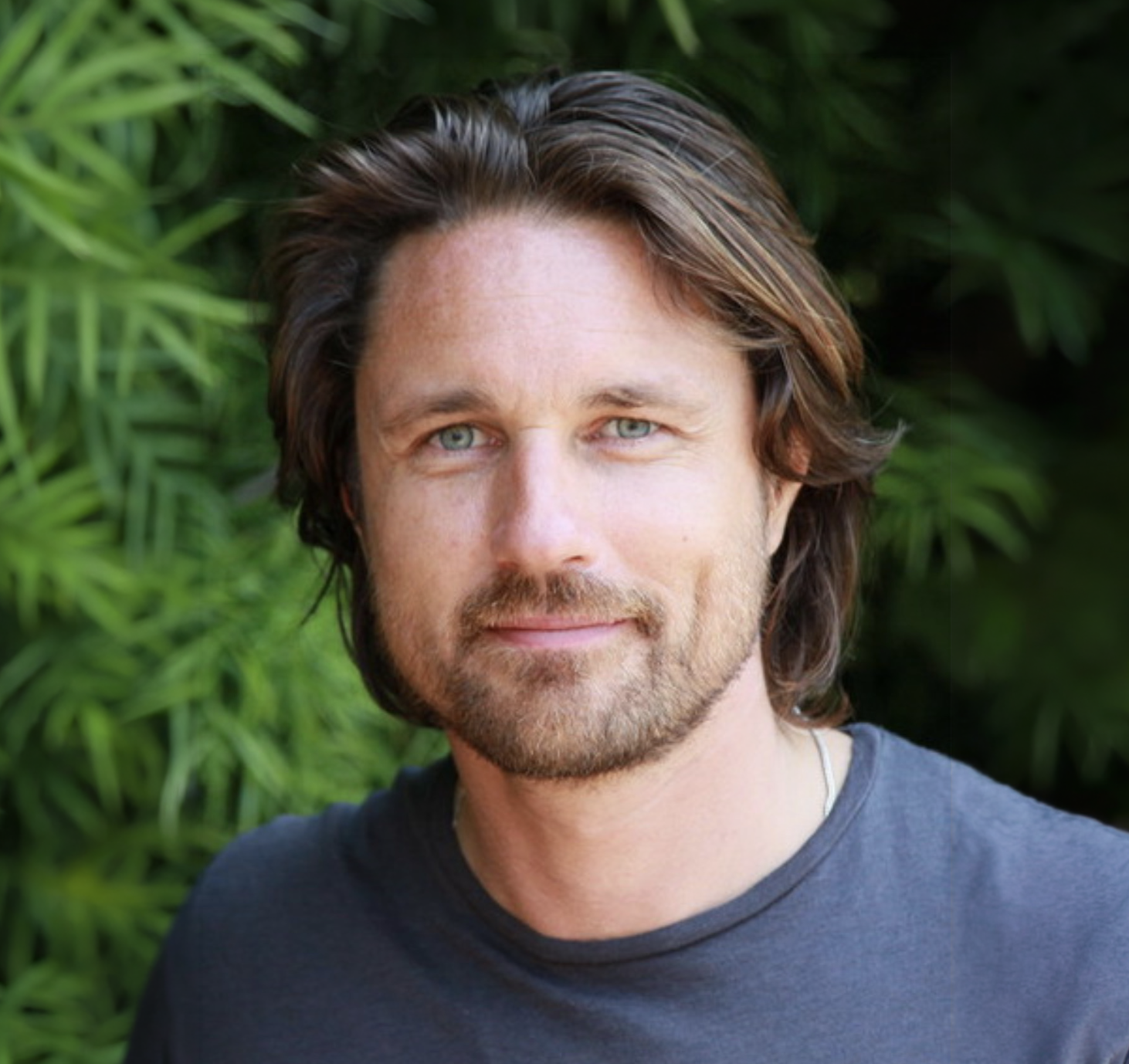 Portrait of a man with wavy brown hair, blue eyes, and a beard, standing outdoors with green foliage in the background.