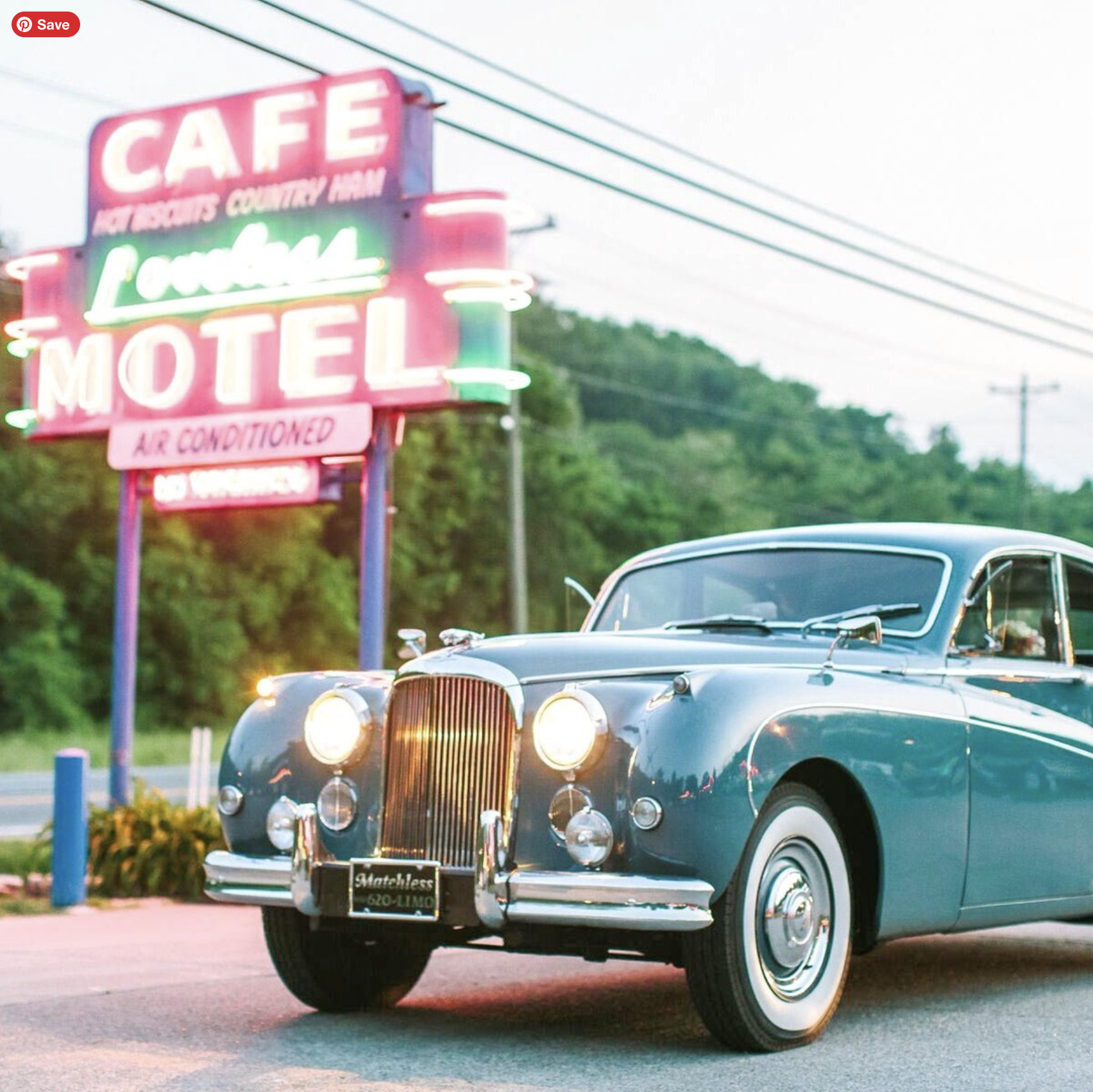 A vintage blue Rolls Royce car parked at a car dealership with a neon-lit motel sign in the background. The sign advertises a cafe, artesian country ham, and air conditioning.