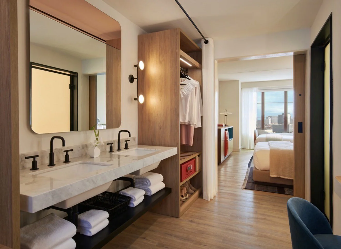 A modern hotel suite with a view of a city skyline through large windows, featuring a bathroom area with a dual sink, wooden cabinetry, and a seating area with a blue chair.