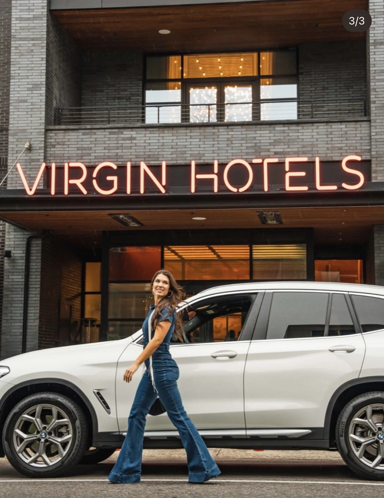A woman in denim jeans and a blue top walking past a white car parked in front of a building with a neon sign that reads 'VIRGIN HOTELS'. The building has large windows and an upper balcony, with indoor lights visible through the windows.