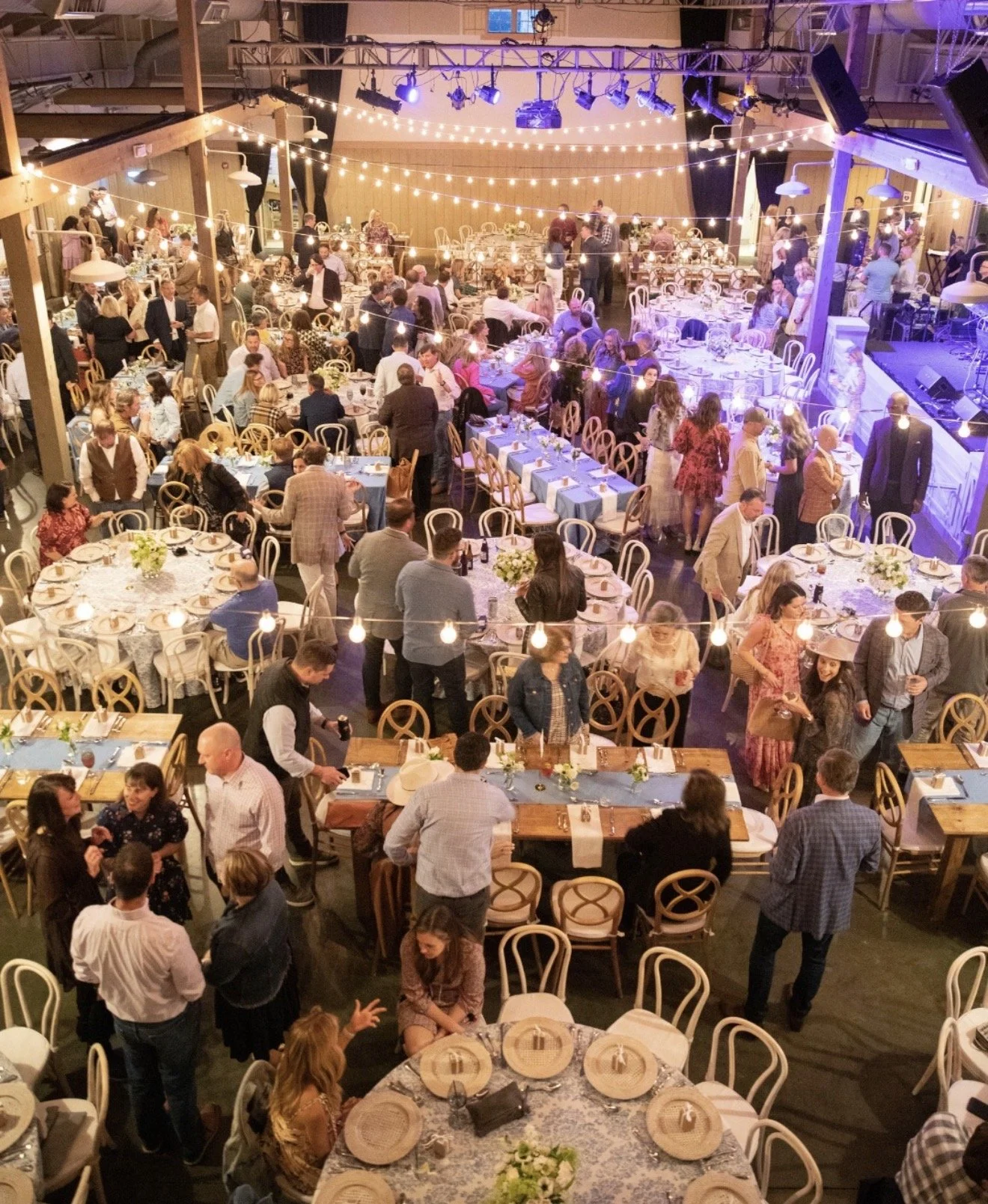 An indoor event with many round tables set for a meal, decorated with flowers and place settings, and surrounded by guests. The space has warm lighting, string lights, and a stage area with purple lighting, with people socializing and mingling.