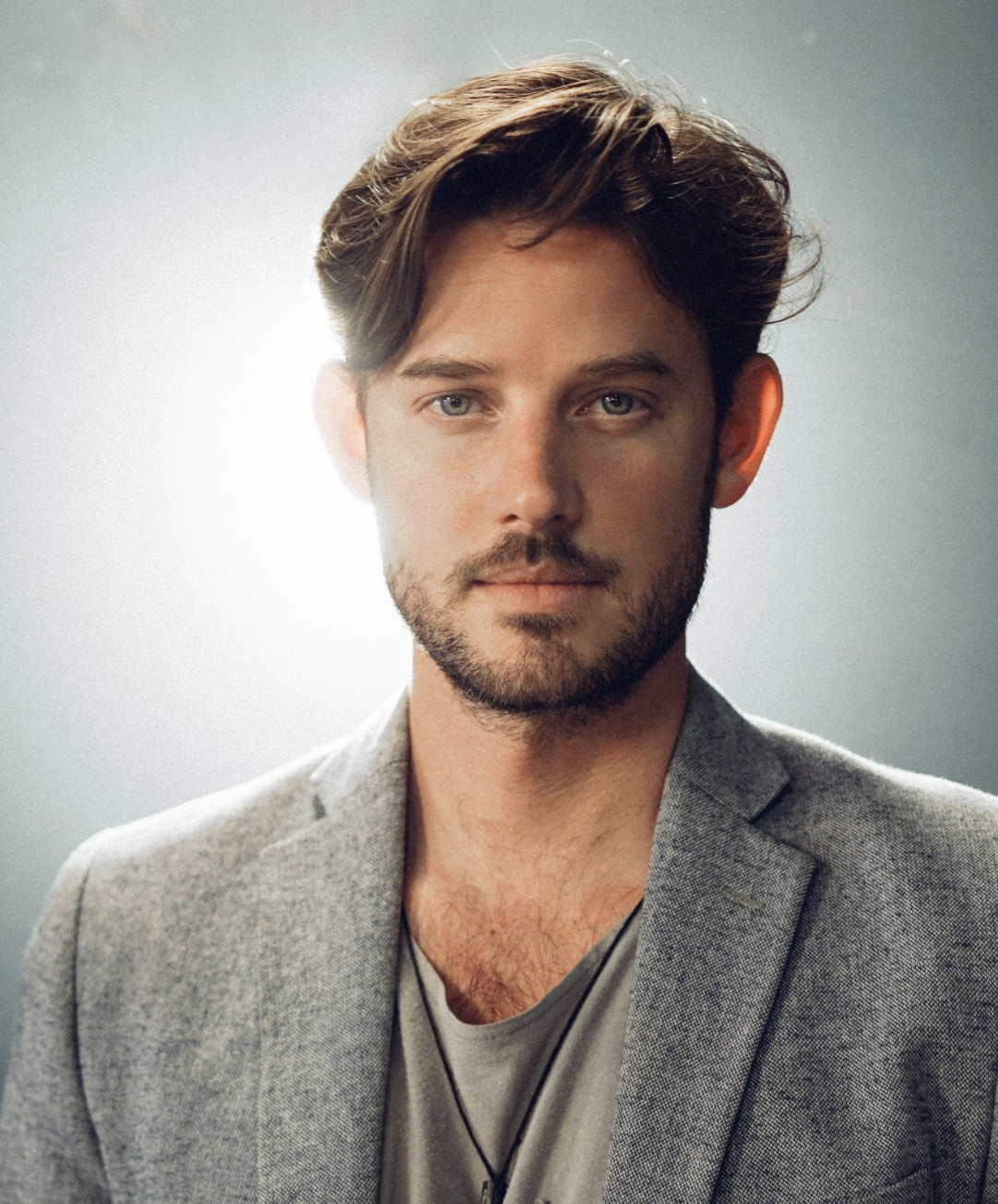 Portrait of a young man with brown hair, beard, wearing a gray blazer and a gray T-shirt, with a neutral expression, against a gray background.