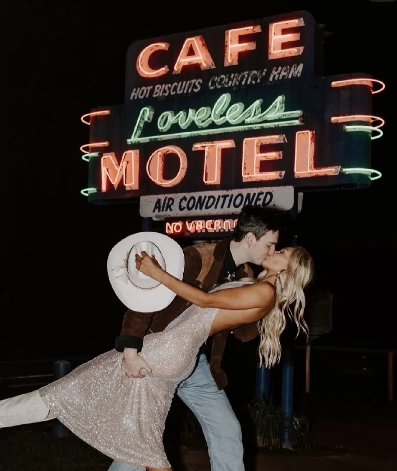 A couple kissing in front of a neon-lit motel sign with a man holding a woman in a glamorous dress while she leans back at an angle.