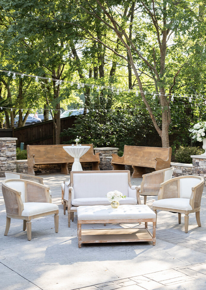 Outdoor patio with cream and wooden seating, string lights overhead, surrounded by lush green trees.