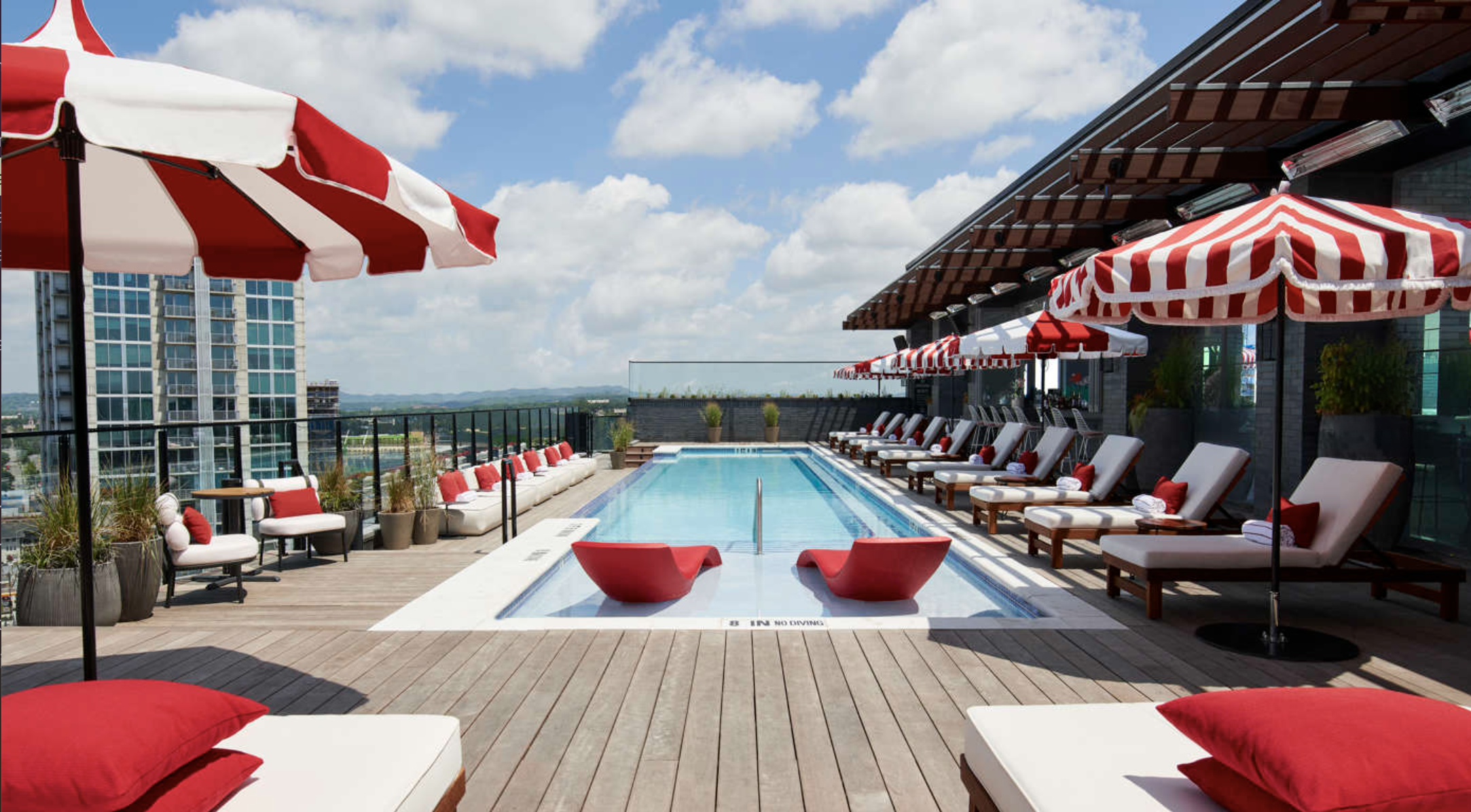 Rooftop pool area with lounge chairs, umbrellas, and city skyline views under partly cloudy sky.