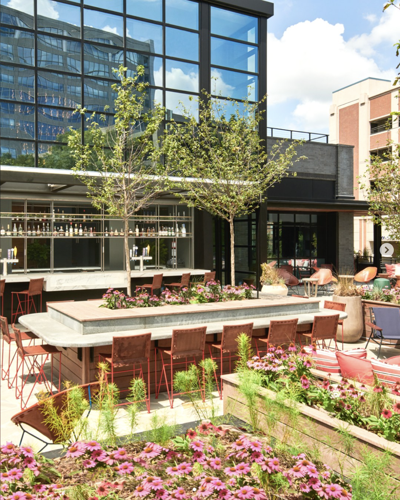 An outdoor patio area with pink flowers, a large rectangular concrete table with pink chairs around it, potted plants, and a modern glass building in the background.