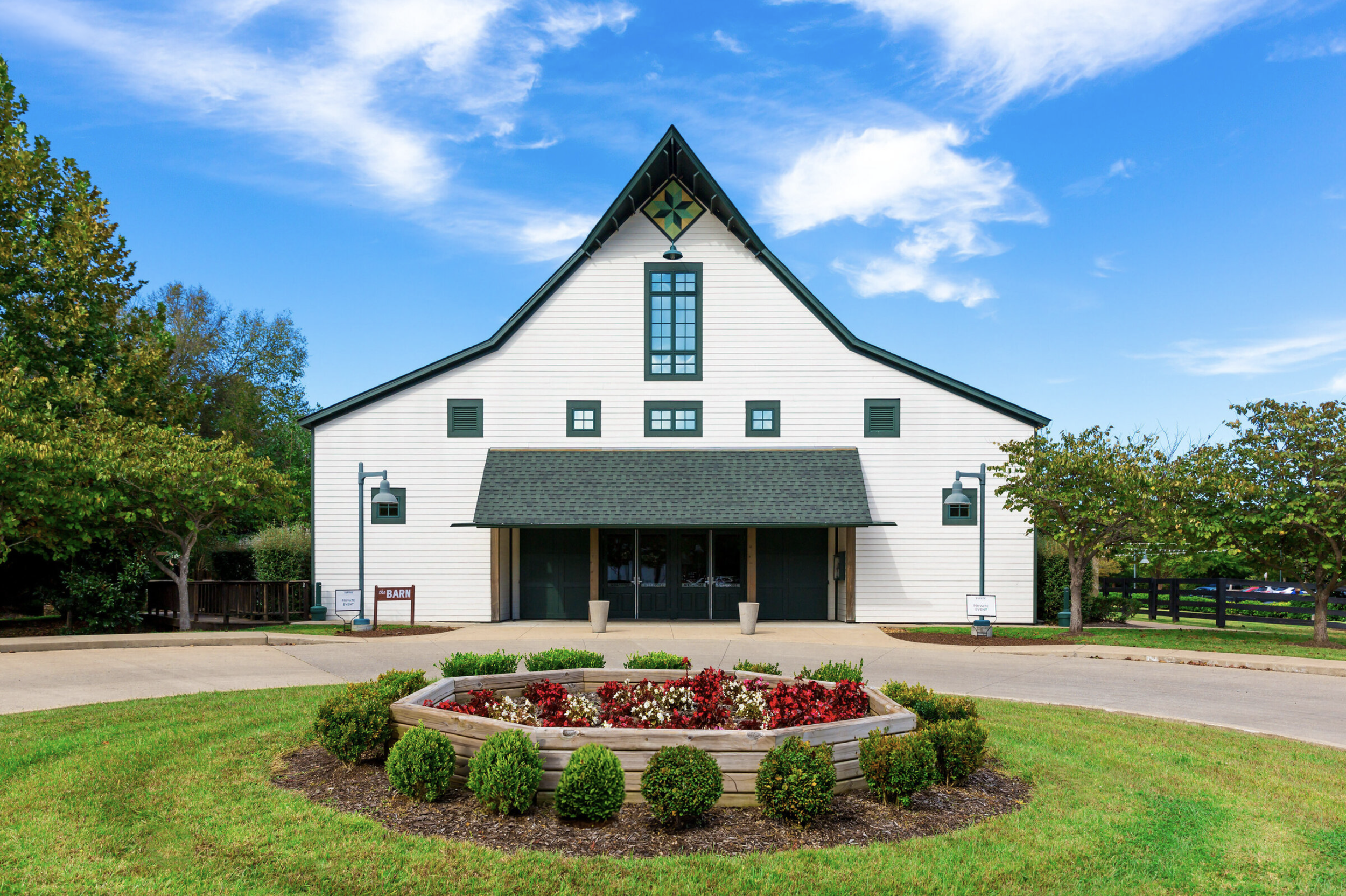 A white barn-style building with a black roof and multiple windows, surrounded by trees and a well-kept lawn with flower beds and signs.