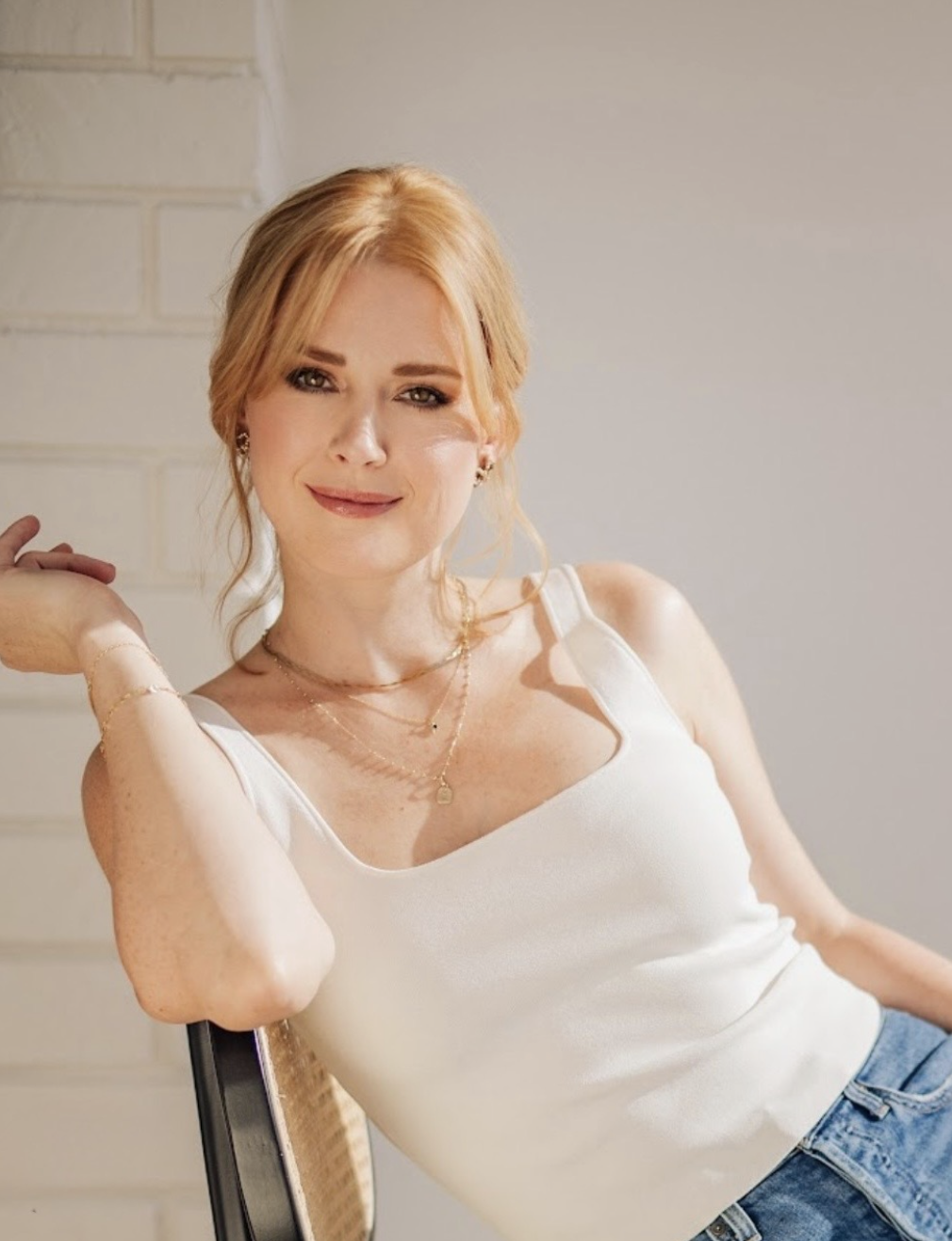 A young woman with red hair, wearing a white top and blue jeans, sitting on a chair with a brick wall in the background, smiling and looking at the camera.