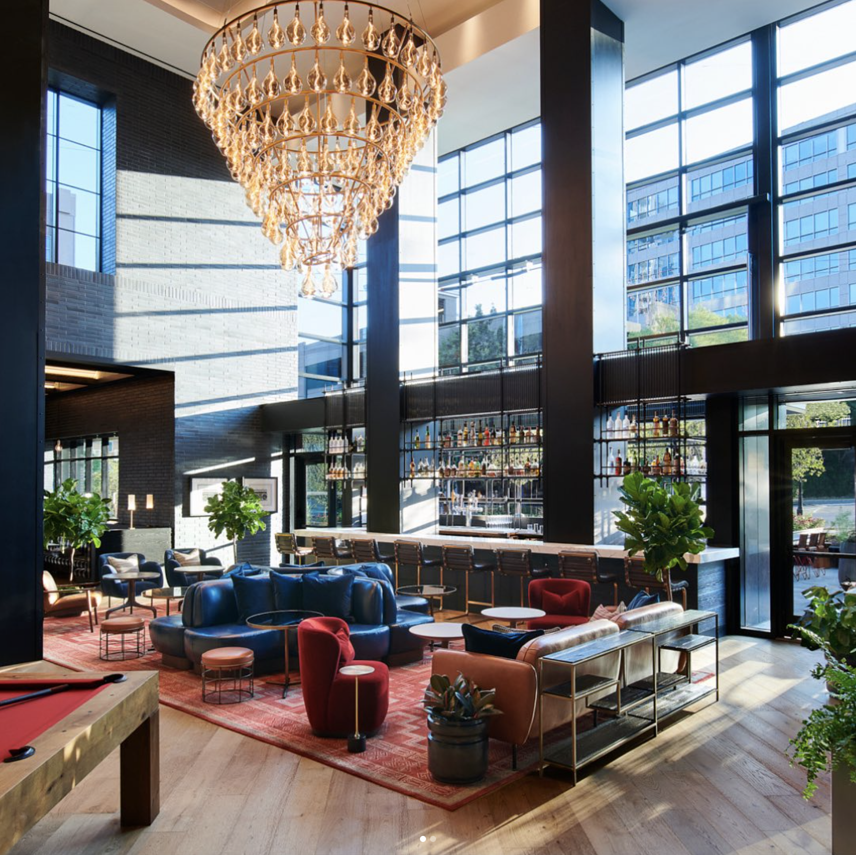 Interior of a modern lobby or lounge area with large floor-to-ceiling windows, a multi-tiered chandelier, contemporary seating, plants, and a bar in the background.