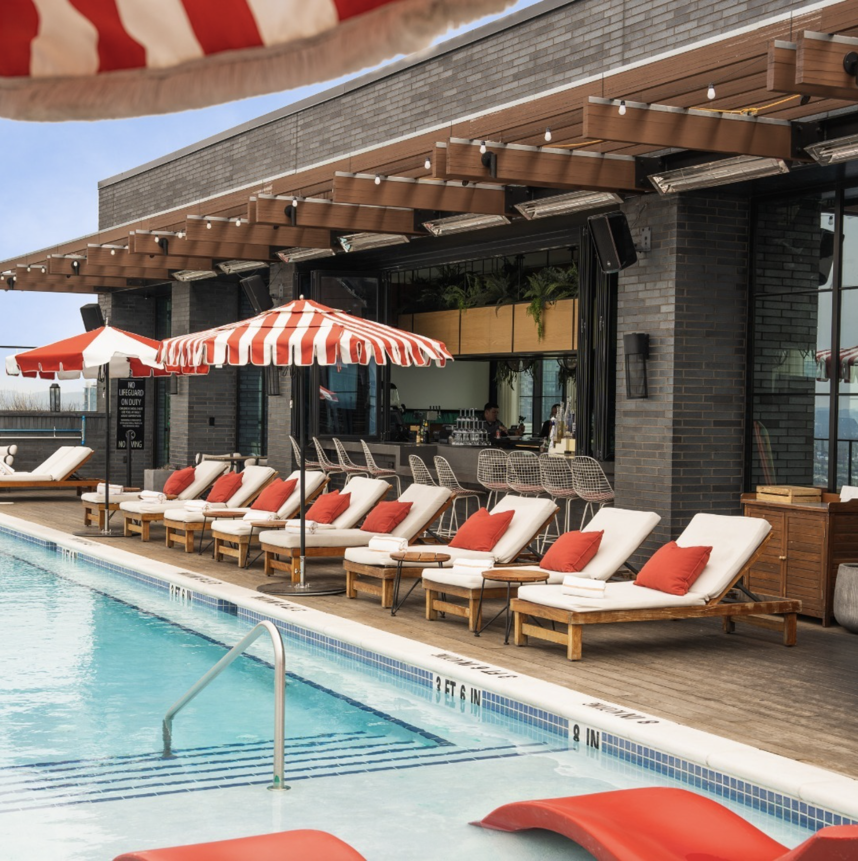 Luxury poolside lounge area with white sun loungers with orange pillows, red-and-white striped umbrellas, and a swimming pool in the foreground. There is a modern building with brick and wood accents in the background, featuring an outdoor bar or seating area with chairs and a bar counter.
