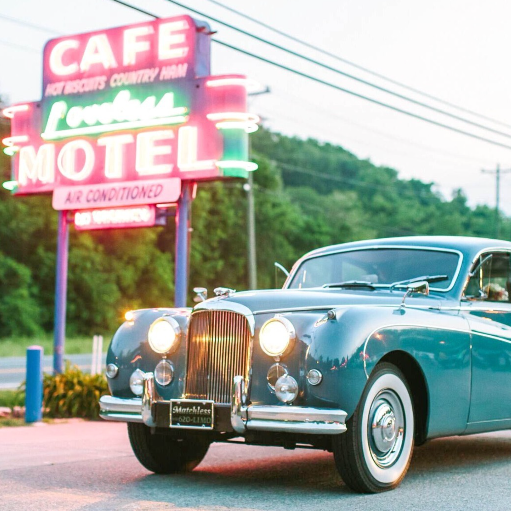 A vintage black Cadillac parked in front of a neon-lit motel sign. The sign reads "Café", "Lounge", "Motel", and "Air Conditioned", with a background of trees and a cloudy sky.