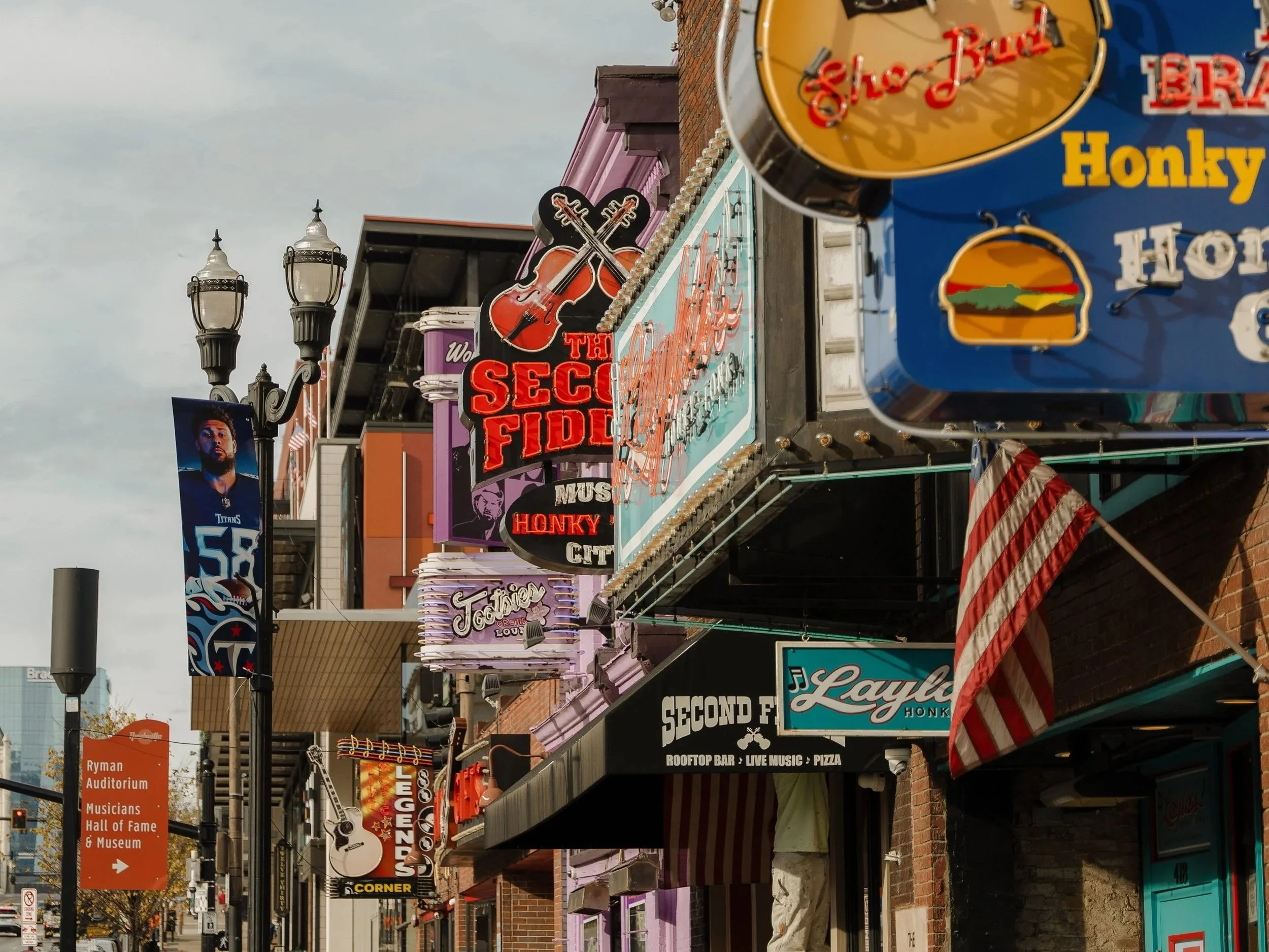 Street scene with various neon signs and storefronts, including signs for a rooftop bar with live music and pizza, a tattoo shop, and entertainment venues, with a large billboard of a football player in the background.