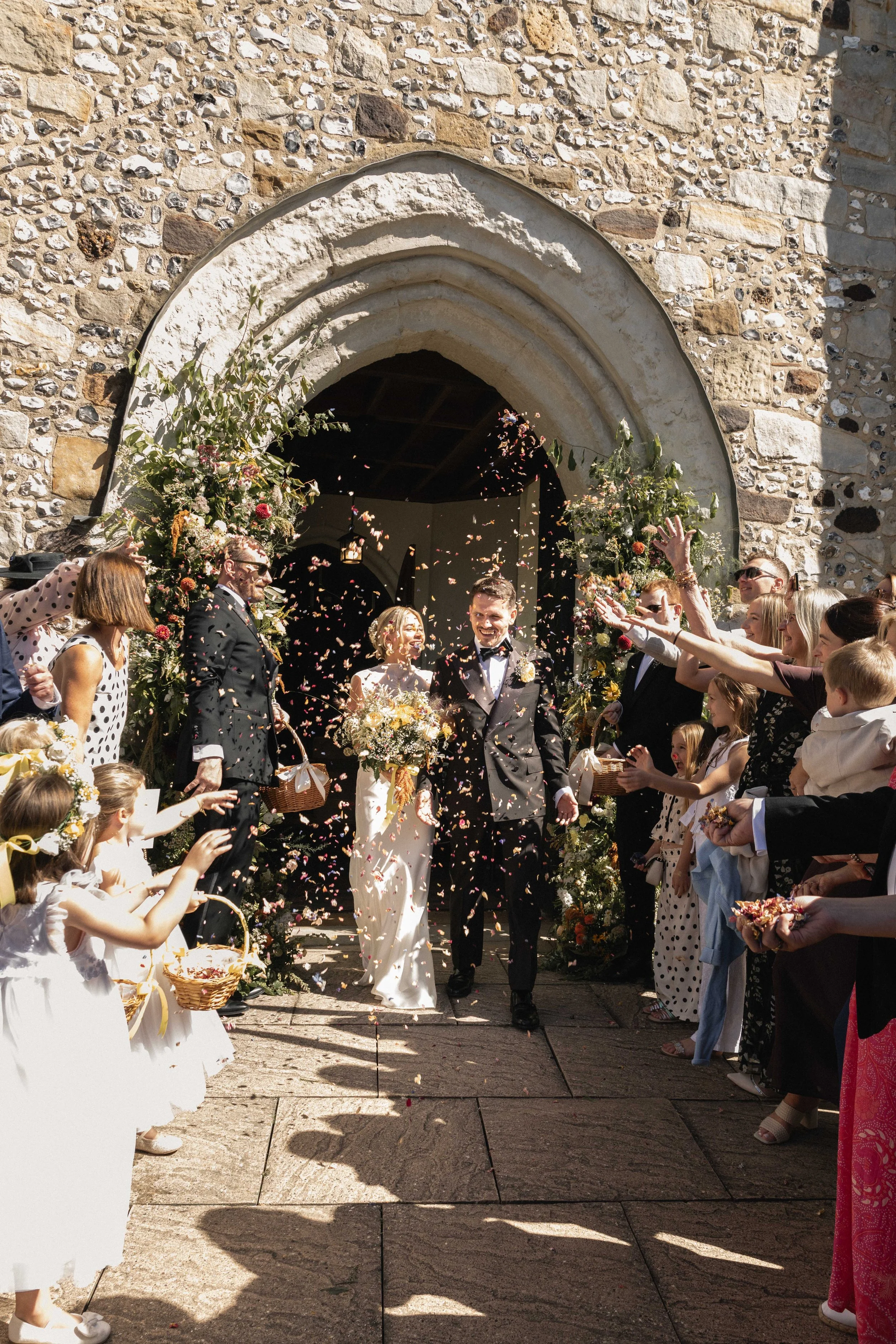 A wedding celebration outside a stone church with guests tossing flower petals at a smiling bride and groom.