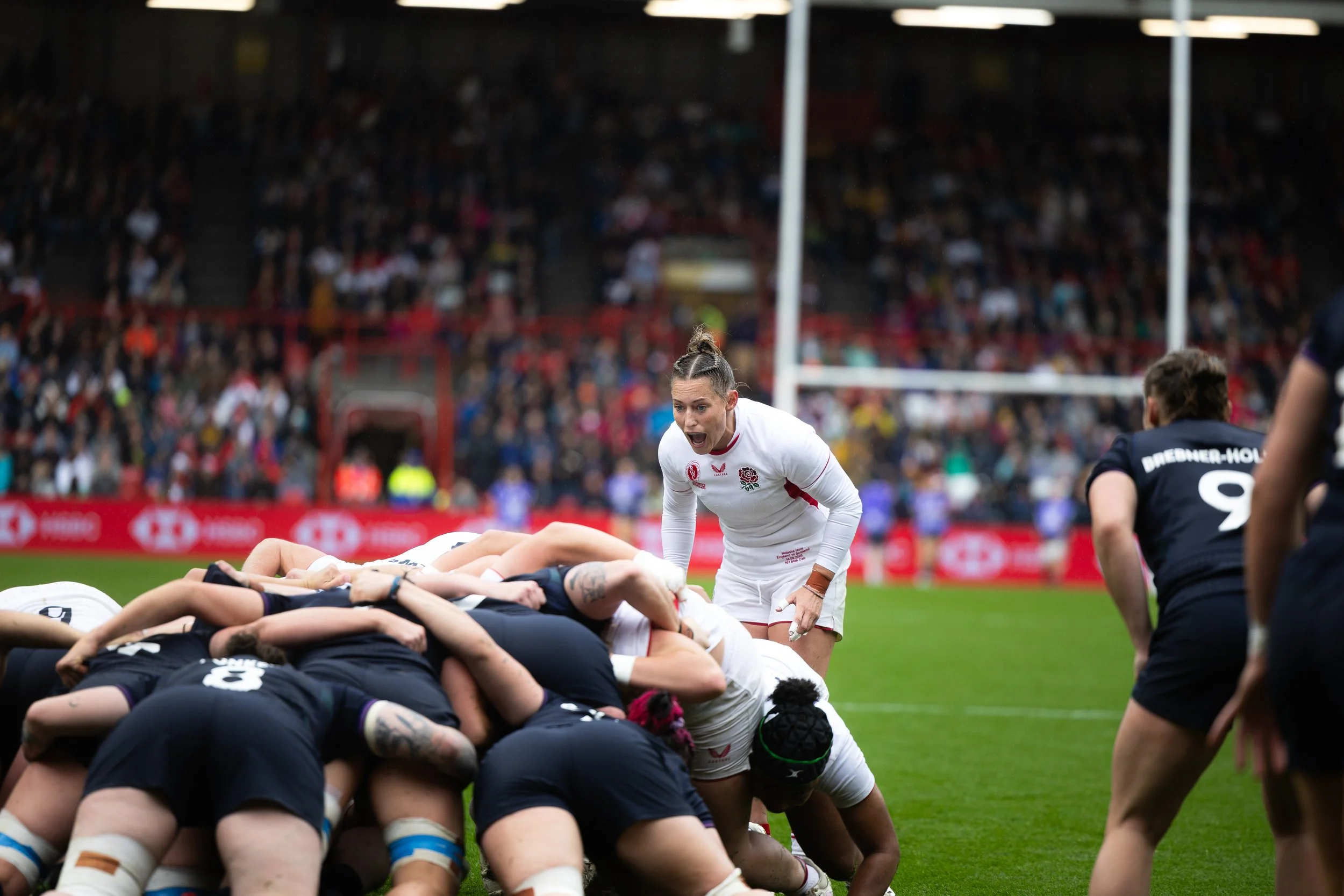 A rugby match with players in a scrum formation on the field, a female player in a white jersey is shouting, with a crowd in the stands.