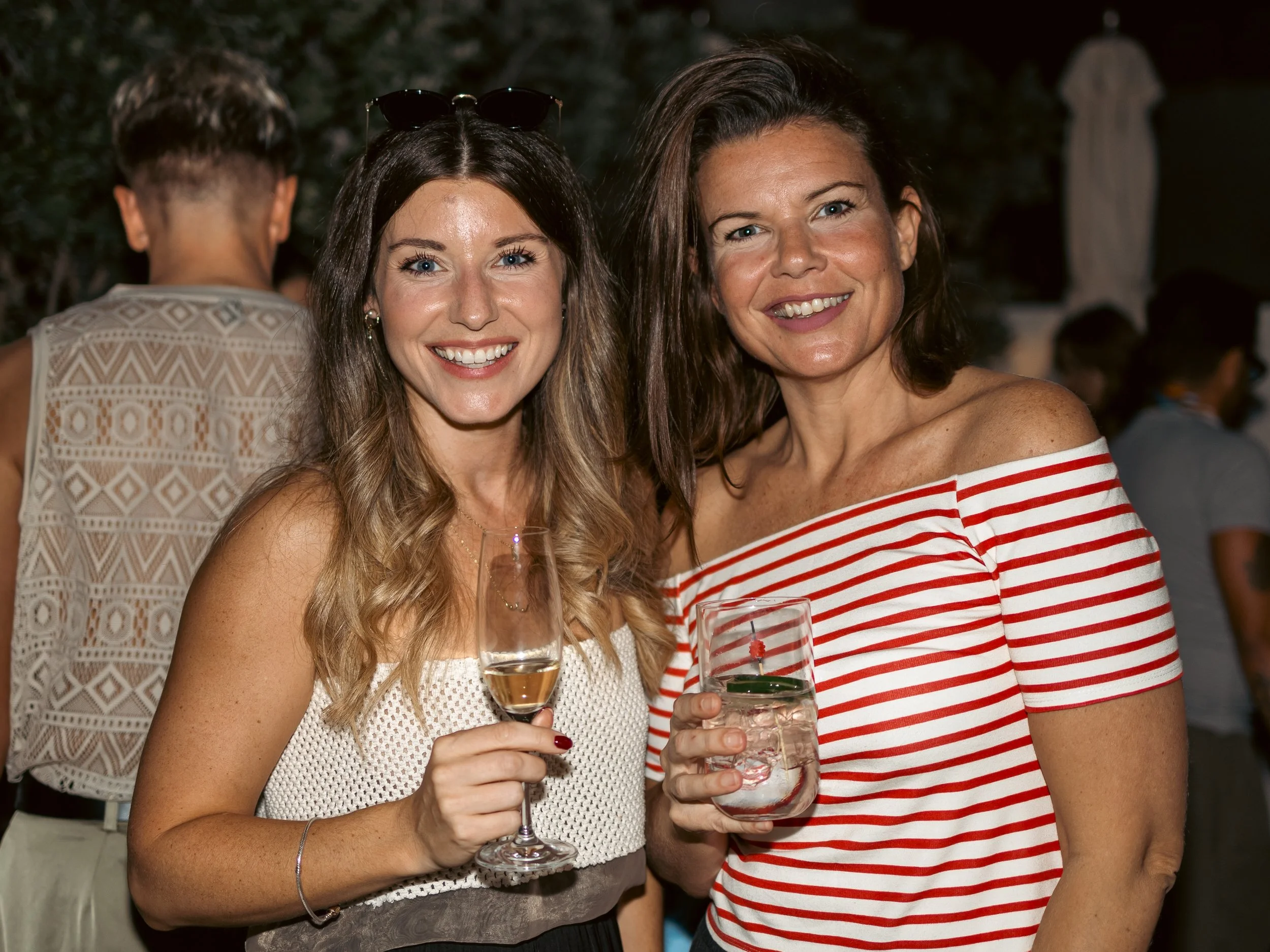 Two women smiling and holding drinks at an outdoor event during the evening. One woman has long, wavy blonde hair and is wearing a white crochet top, while the other has shoulder-length dark hair and is wearing a red and white striped top. They are s