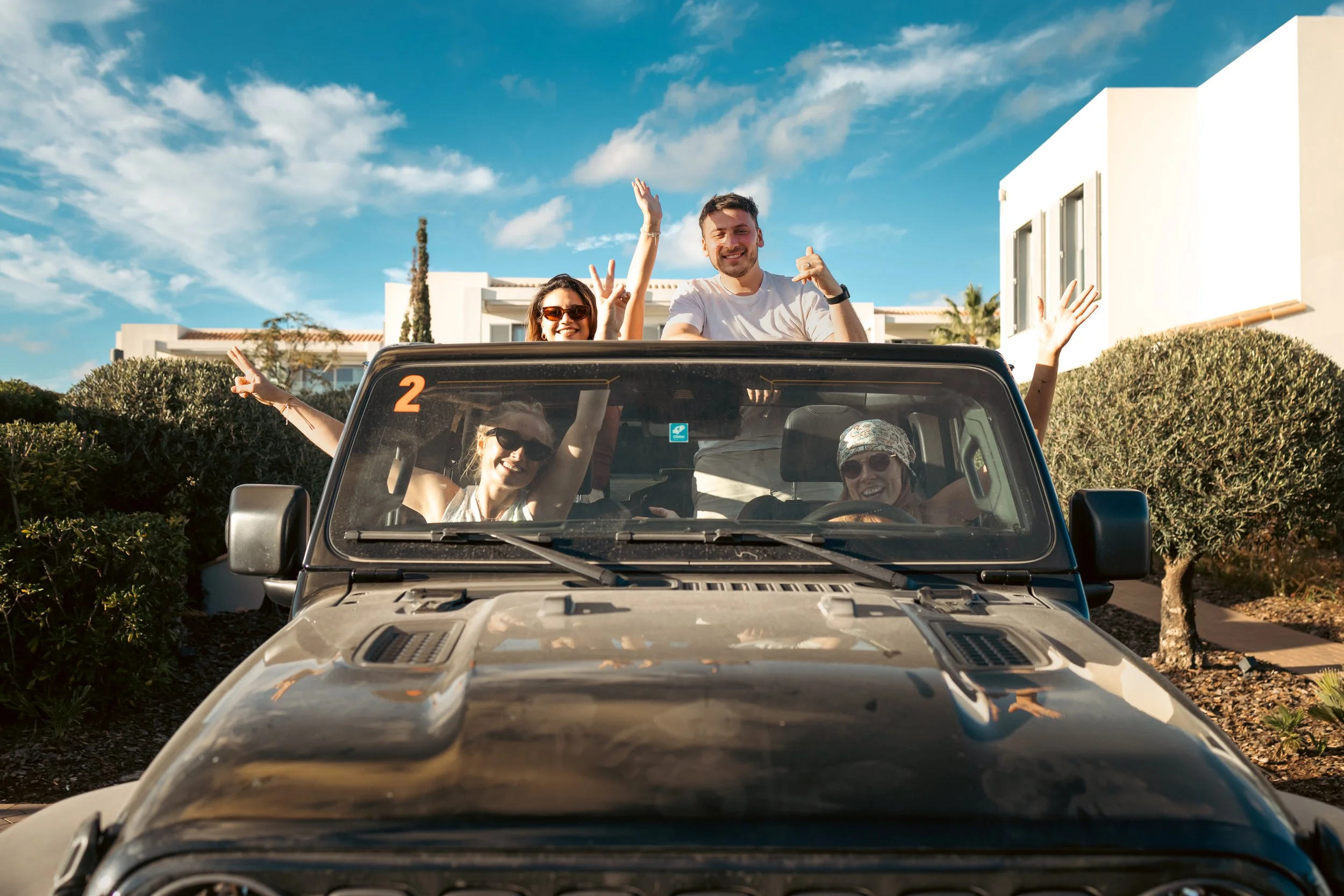 People enjoying a sunny day in a black convertible car, with two women in the front and two men waving from the back, surrounded by greenery and modern white buildings, under a blue sky with some clouds.