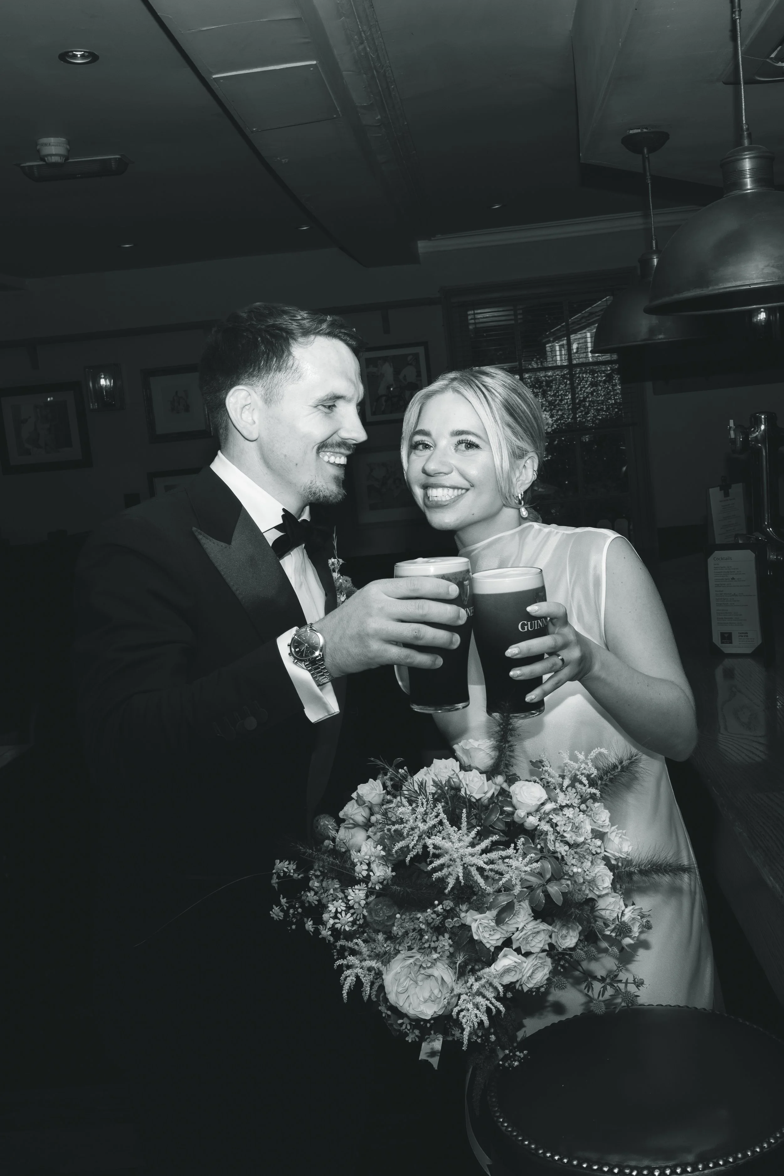 A happy bride and groom at their wedding celebration, holding glasses of dark beer, with a bouquet of flowers in front of them, smiling and clinking glasses.
