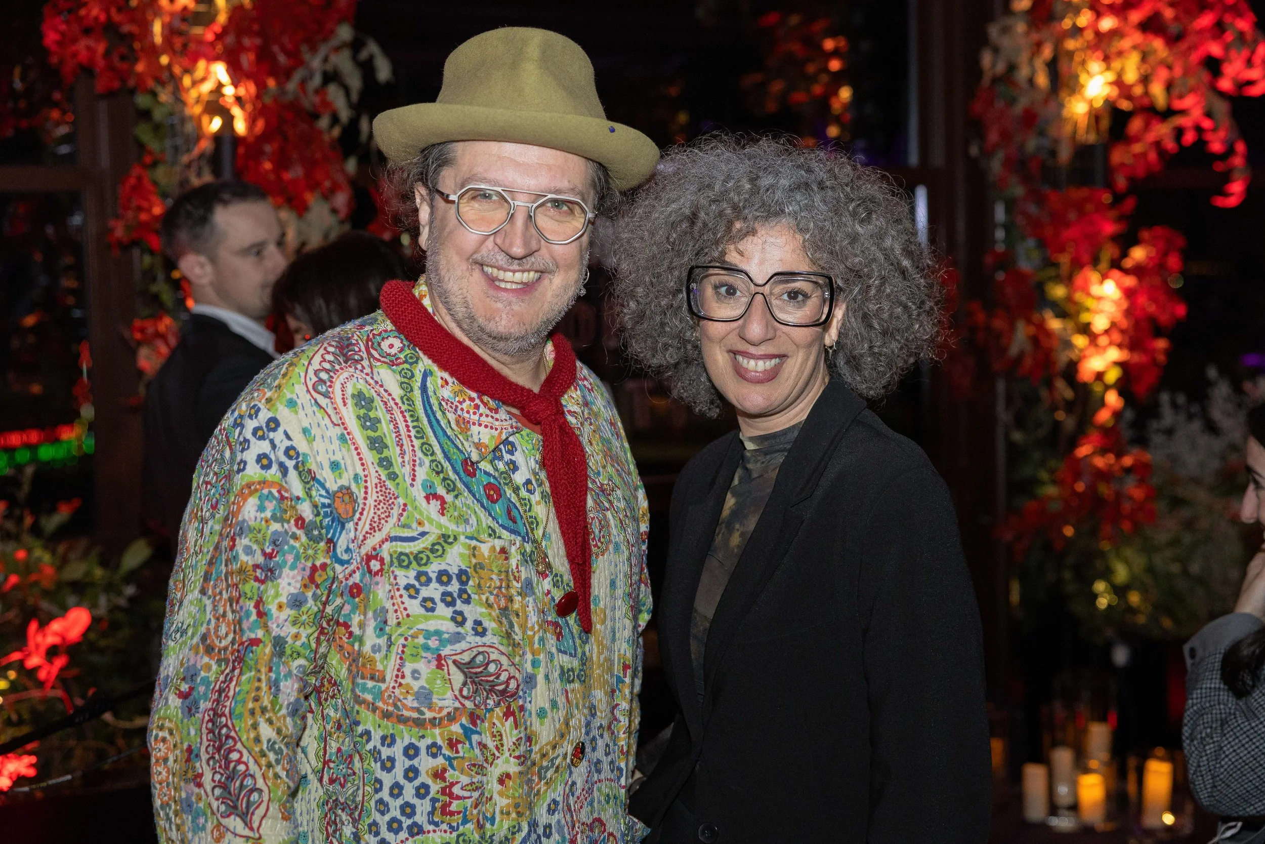 Two smiling people standing close together at an outdoor event, with people and vibrant red and orange foliage in the background.