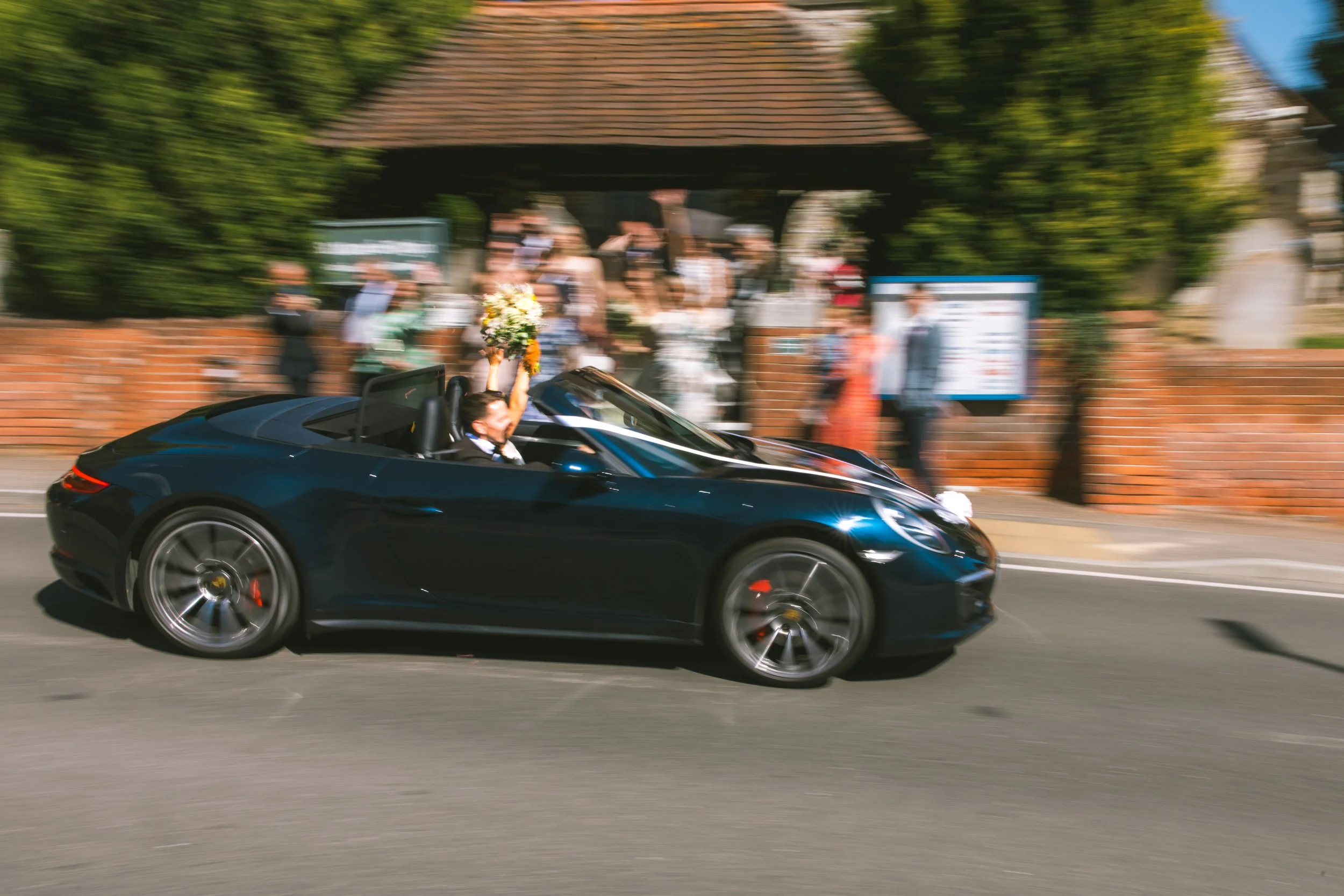 A black convertible sports car driving past a park with people gathered near a brick wall and trees. The driver is holding a bouquet of flowers.