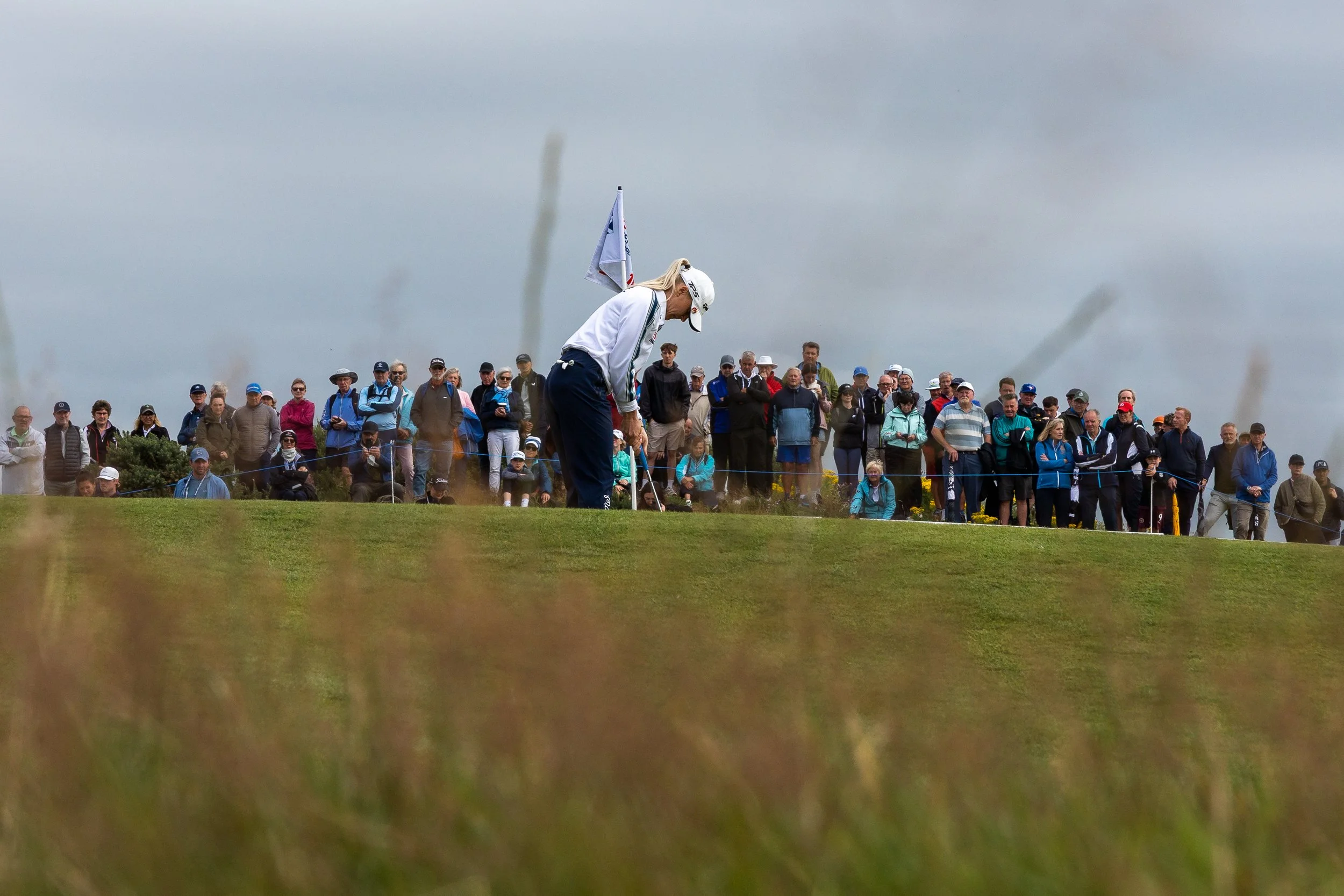A female golfer practicing her shot on a grassy golf course with a group of spectators watching, overcast sky.