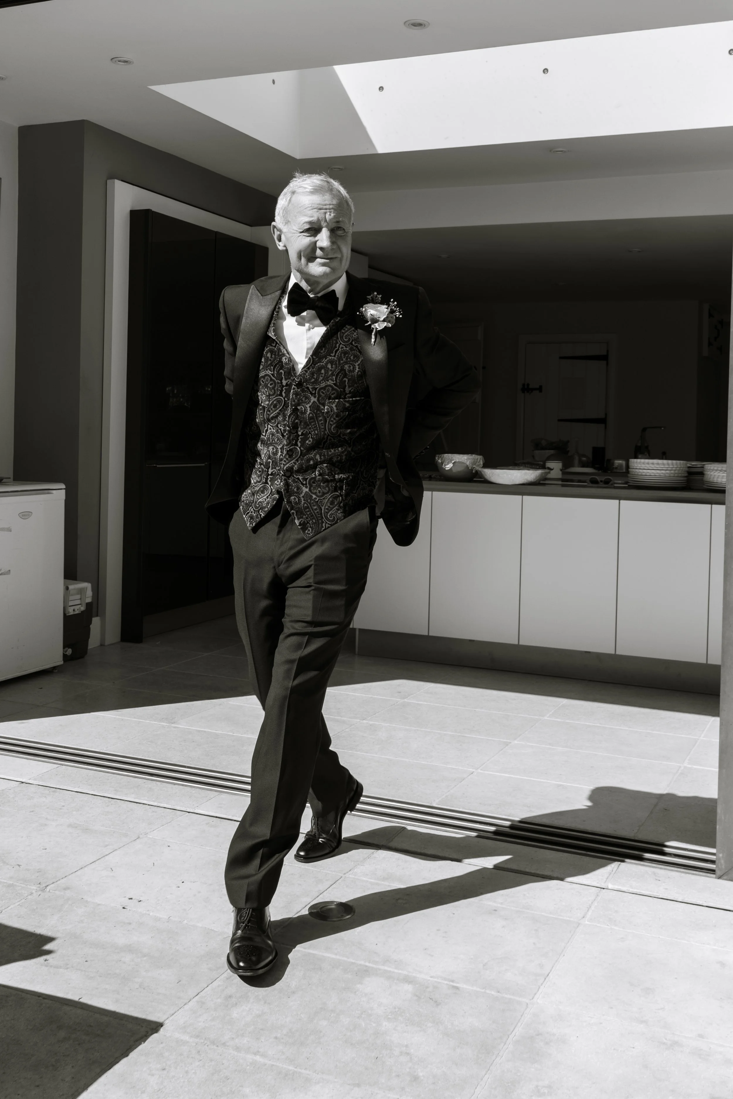 A man wearing a tuxedo with a bow tie and boutonniere stands outdoors in sunlight, with one arm behind his back and one leg crossed in front of the other, on a patio area with modern kitchen in the background.